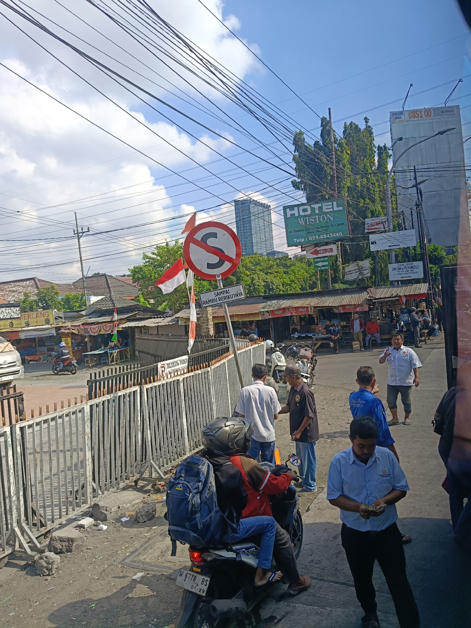 The image captures a street scene in Kedungrejo, Indonesia, under a bright, cloudy sky. In the foreground, a white fence runs along the left side, separating a sidewalk from the street. Several people are visible, some standing and interacting, while others are on or near motorcycles. One man in a light blue shirt is holding and counting money. A motorcycle with a blue backpack strapped to the back is parked near the fence.
Further back, there are several small food stalls or shops with awnings. Indonesian flags are displayed on poles in front of some of these establishments. A "no U-turn" traffic sign is prominently positioned on the right side of the image, accompanied by Indonesian text on a white sign.
In the background, several buildings are visible, including a multi-story modern building and a larger structure with "HOTEL WISTON" signage. Power lines crisscross the sky above the scene. The overall impression is of a bustling commercial street with a mix of everyday activities and traffic control measures.