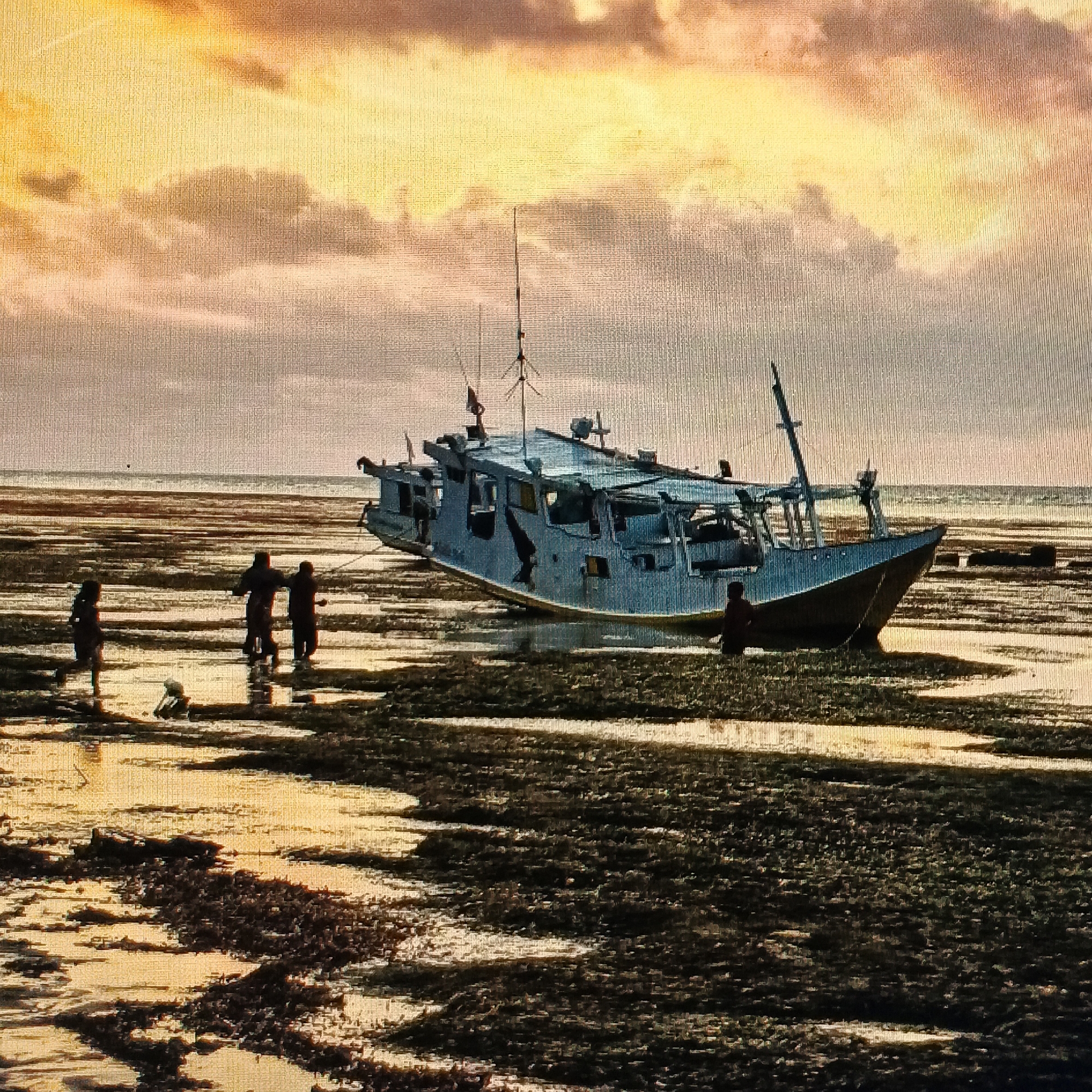 The image depicts a fishing boat aground on a muddy tidal flat during low tide, with several individuals present. The boat, a light blue vessel with a yellow hull, is positioned horizontally across the scene, angled slightly away from the viewer. It appears to be a traditional fishing boat with a superstructure and masts. The surrounding environment is characterized by the exposed seabed, covered in dark mud, sand, and seaweed, with shallow pools of water reflecting the sky.

In the foreground and to the left of the boat, three figures are visible. One figure, in the far left, appears to be walking or standing. Two figures are standing together further to the right, one slightly behind the other. To the right of the boat, a single figure is standing, facing away from the viewer. The figures are silhouetted and their features are indistinct.

The sky is filled with dramatic clouds, illuminated by what appears to be sunset or sunrise, casting a warm, golden-orange glow over the scene. The light creates reflections on the wet surfaces of the tidal flat. The overall impression is one of a rural coastal setting at dusk or dawn.

While the specific location within Ende, Indonesia, is not explicitly identifiable from the image alone, such scenes are common in coastal areas like the ones found in and around Ende, where fishing is a primary activity and tidal flats are characteristic of the coastline.