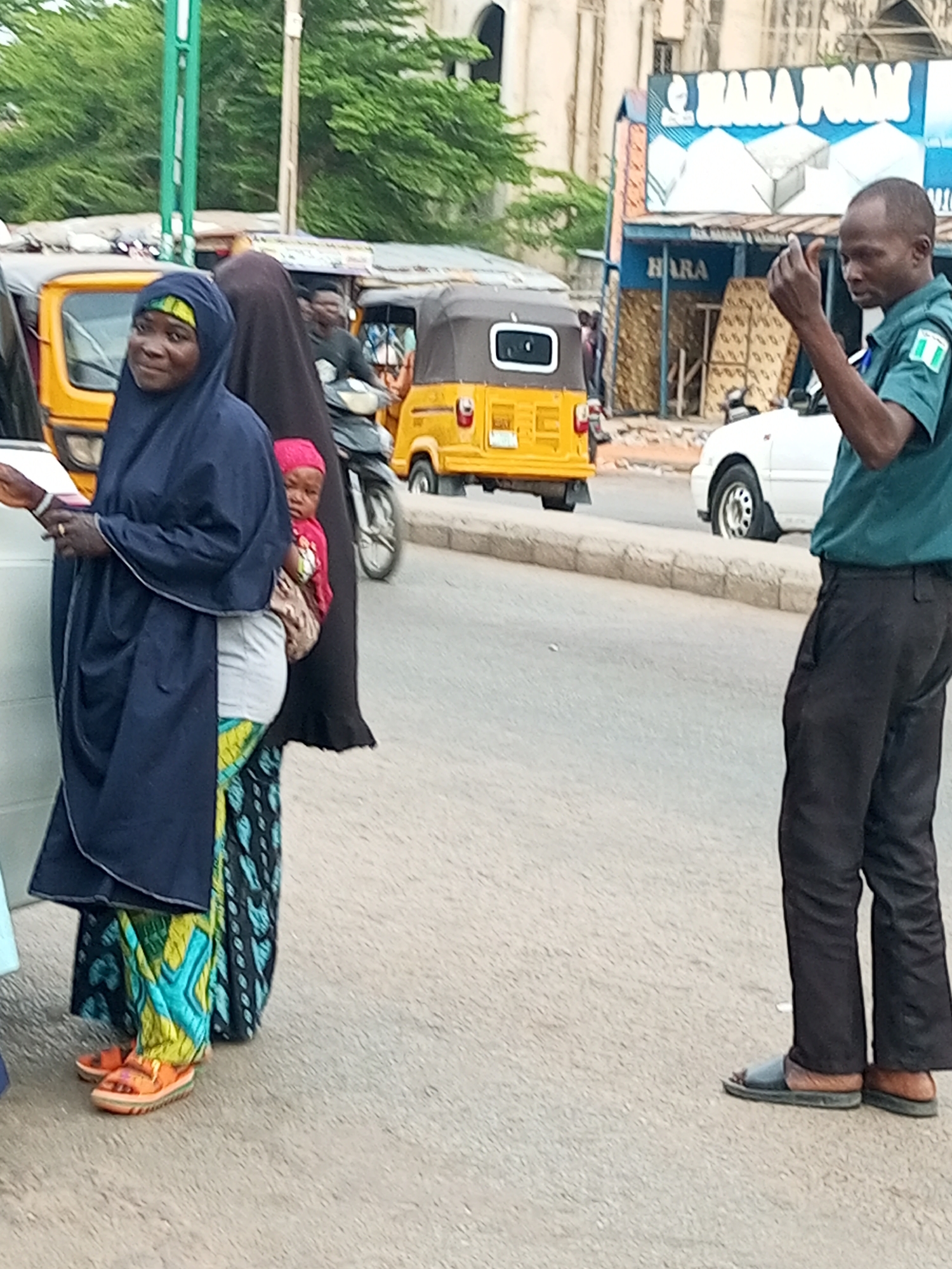 The image captures a street scene in Arawa, Nigeria. In the foreground, a woman wearing a dark blue hijab and a vibrant, patterned skirt is standing, holding a small child in a pink garment. She appears to be looking towards the left, possibly at someone or something off-camera. To the right of the woman, a man in a green uniform with a Nigerian flag emblem on his sleeve is visible. He is looking to his right and holding up his hand, appearing to gesture or communicate. His dark pants and open-toed sandals are also visible.
Behind the individuals, several vehicles are present. A yellow auto-rickshaw, commonly known as a "Keke Napep," is prominently parked on the left. Further back, another similar vehicle and a white sedan are visible. The background features buildings, one of which has a large blue and white sign that reads "HARA FOAM" and also "HARA." There are also trees with green foliage, suggesting it might be daytime with moderate to warm weather. The overall atmosphere suggests a busy street or market area, with people going about their daily activities. The people are dressed in a way that is common for the region.