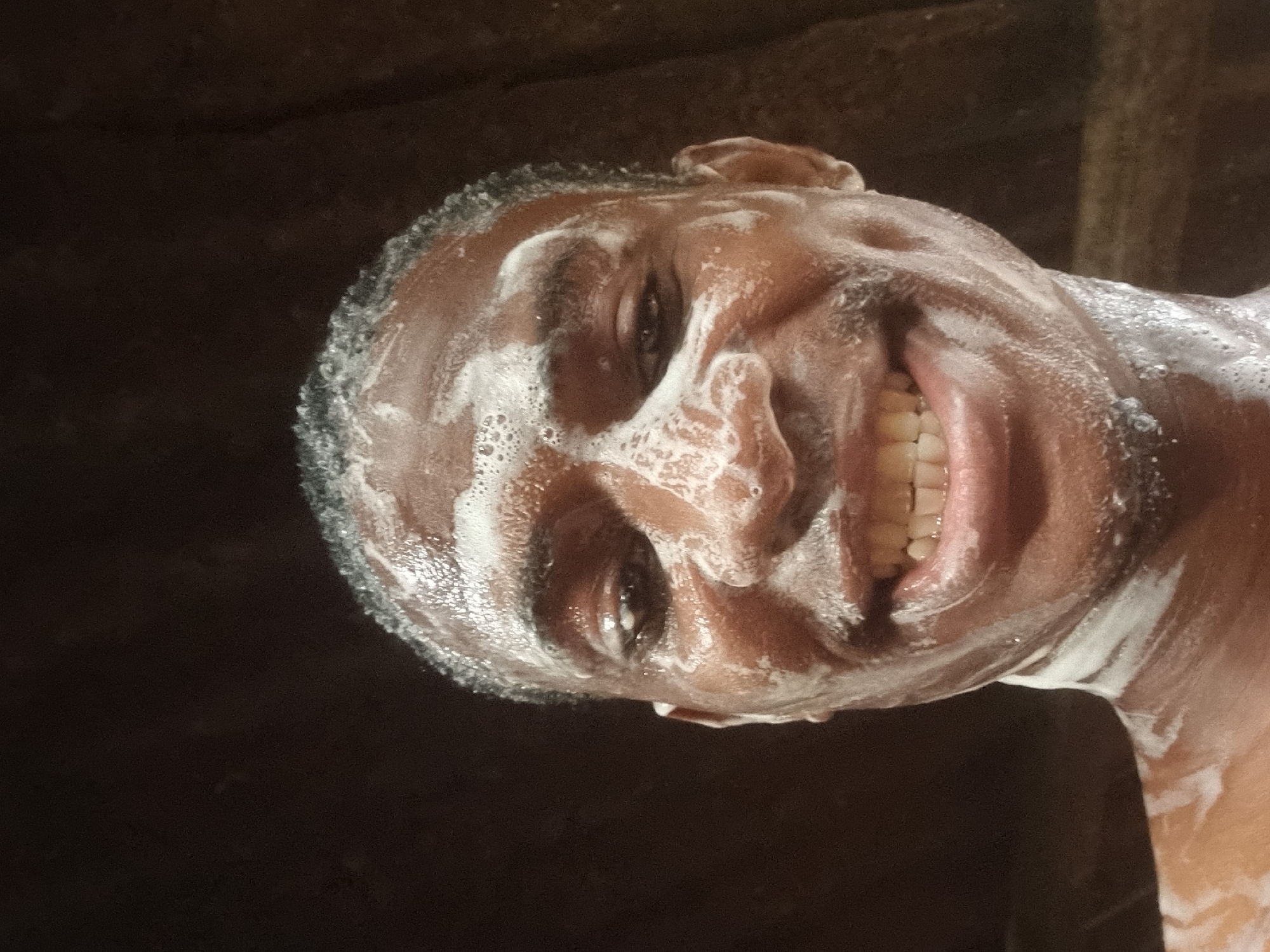 A dark-skinned man is seen from a low angle, presumably in a bathroom setting. Soap covers his head and parts of his face. He is smiling, exposing his teeth. The background is dark. There is no indication that the image was taken in Onitsha, Nigeria.