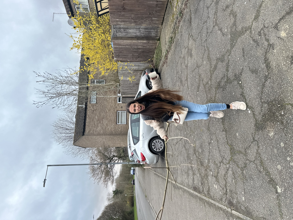 The image depicts an outdoor scene on an overcast day in Corby, United Kingdom. A female subject runs or strides towards the camera on an asphalt surface. She has long, dark brown hair and is dressed in a light-colored fuzzy jacket over a dark top, light blue skinny jeans, and light-colored sneakers. Her mouth is open, and her arms are outstretched. In her left hand, she holds a long, slender, multi-branched wooden stick. Her right hand grips a white disposable cup with a dark lid. A small, light-colored shoulder bag with a distinct emblem is worn across her body.
The asphalt ground displays numerous cracks and signs of wear. To the left of the subject, a curb delineates the pavement from a road. Several silver hatchback cars are parked along the road. A grey street lamp post stands on the left. In the background, a two-story brick residential building with a dark roof and a satellite dish is visible behind a dark brown wooden fence. Bare deciduous trees are present to the left and behind the building, while a tree or bush with bright yellow blossoms is visible behind the fence to the right. The sky is uniformly overcast.
