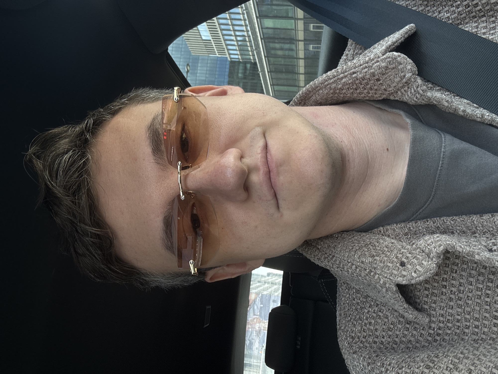 A selfie captures a person wearing rimless, orange-tinted glasses, likely seated in a car given the presence of a seatbelt. The person wears a light gray shirt and a tan jacket. The background shows a glimpse of the cityscape with modern office buildings, suggesting an urban environment.