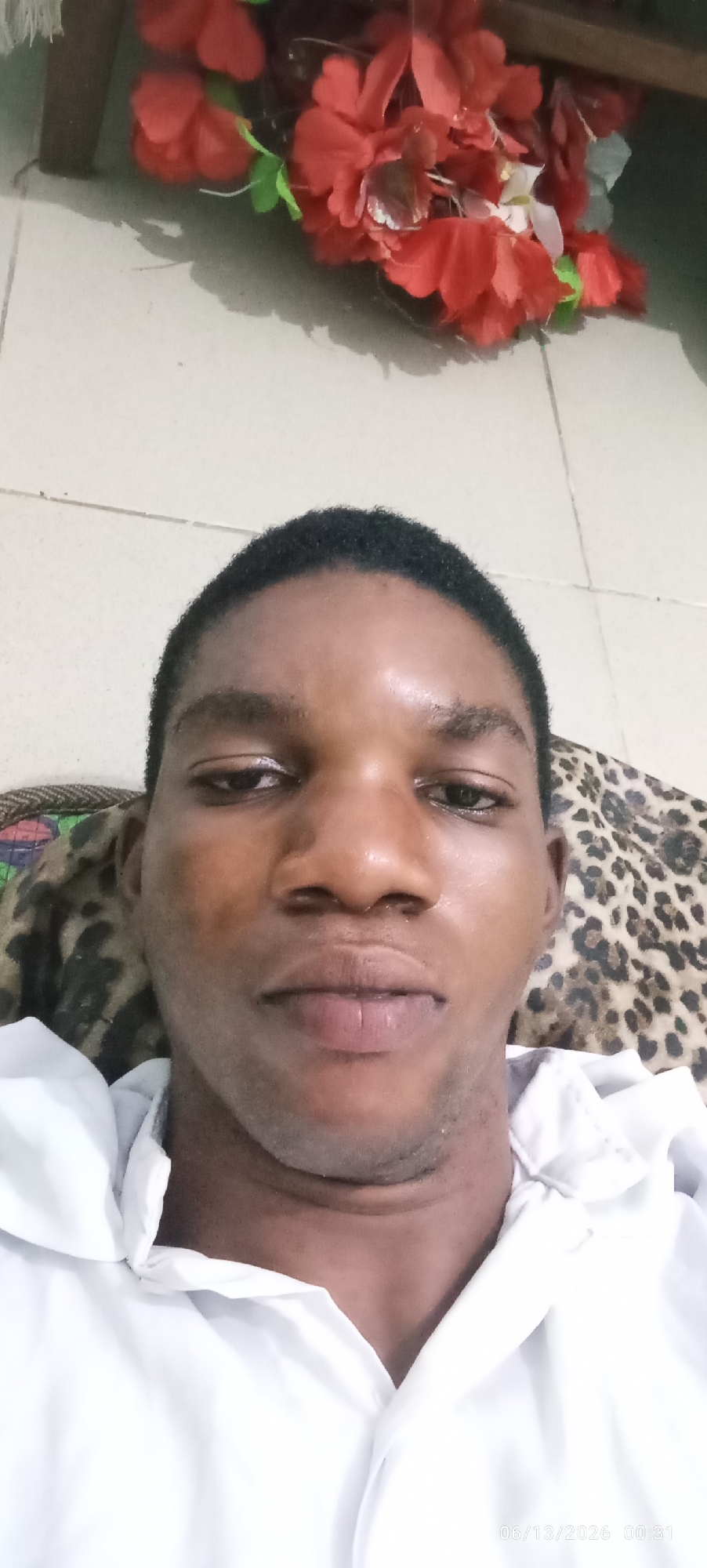 A close-up, eye-level, selfie-style photograph features a young Black man looking directly at the camera. He has short, dark hair and a dark complexion. He is wearing a white collared shirt. Behind him, to the left, is a glimpse of what appears to be a woven material with a patterned design. To the right and slightly behind him is a leopard-print fabric. In the upper background, out of focus, are bright red artificial flowers. The floor appears to be tiled in a light color. The lighting suggests an indoor setting, likely at night given the timestamp in the lower right corner: "06/13/2026 00:31". The image is centrally focused on the man's face, with the background elements providing context but not dominating the frame. It is not possible to determine the specific location in Lagos, Nigeria from the visual information provided.