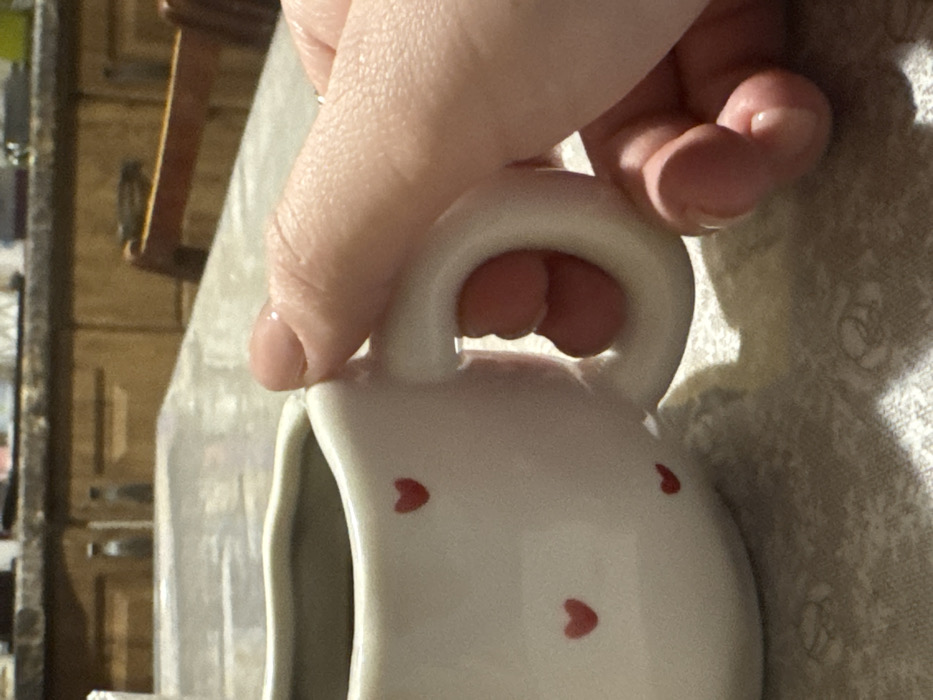 A close-up photograph captures a light-skinned human hand, exhibiting short, clear-polished fingernails, firmly gripping the handle of a white ceramic creamer or small pitcher. The vessel is adorned with multiple small, red heart-shaped decals. This scene is situated on a table covered by a light-toned, possibly off-white, patterned tablecloth. The background, out of focus, reveals wooden kitchen cabinetry with visible handles and a portion of a dark-stained wooden chair, indicative of a domestic kitchen or dining environment in Overyata, Russia. The action depicted is the manual grasping of the creamer.