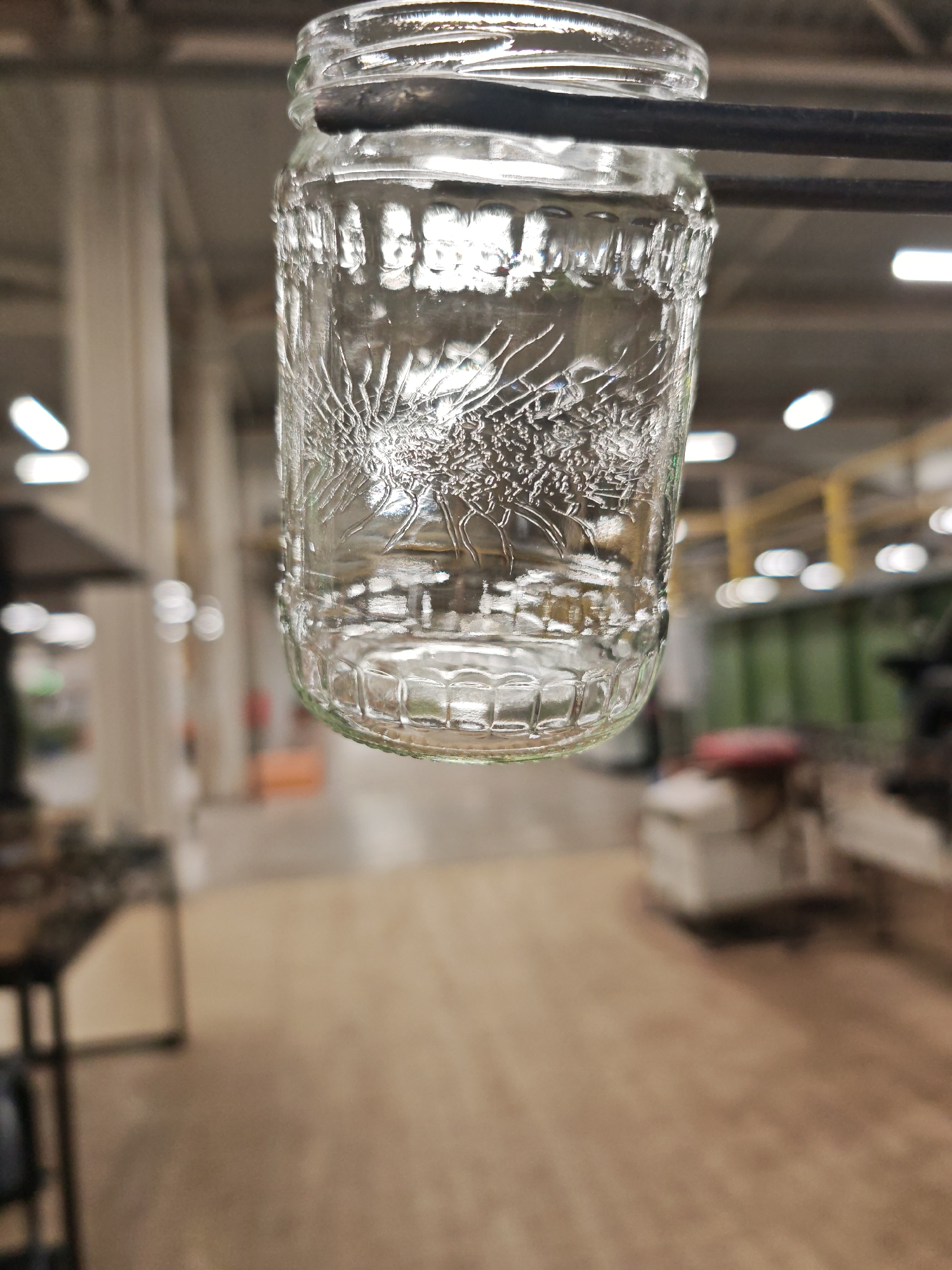 A glass jar is suspended from a metal rod in an industrial setting. The jar has a decorative design etched into its surface, possibly a tree or floral motif. The background is blurred, showing industrial equipment and lighting. The location is potentially a factory or workshop in Pleven, Bulgaria, but this is not definitively confirmed by the image alone.