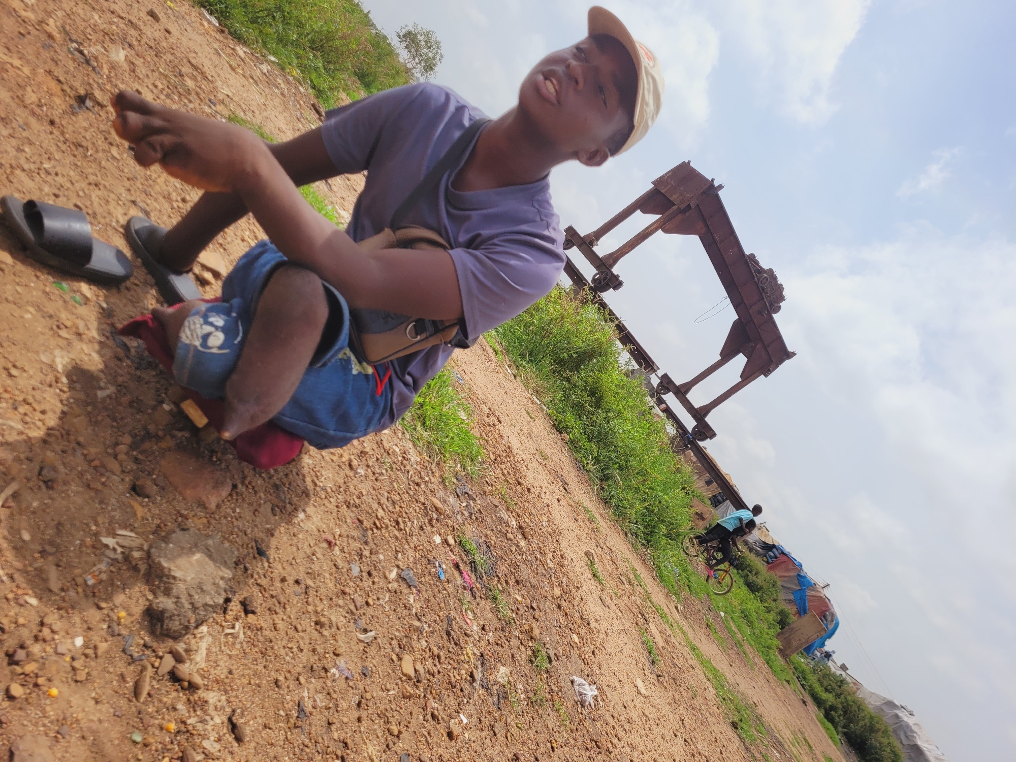A young Black male is crouching on a gravelly surface in Bukuru, Nigeria, under a partially cloudy sky. He is wearing a purple t-shirt, blue shorts adorned with a skull patch, and a tan baseball cap. A small brown bag is slung across his chest. His right hand is extended towards the ground. Two black flip-flops are visible near his feet, one on his left foot and another lying on the ground. In the background, a large, rusted metal structure, possibly a gantry crane, dominates the horizon. Further in the distance, several temporary structures covered with tarps and a person riding a bicycle can be seen. The ground is a mix of dirt, small stones, and scattered debris.