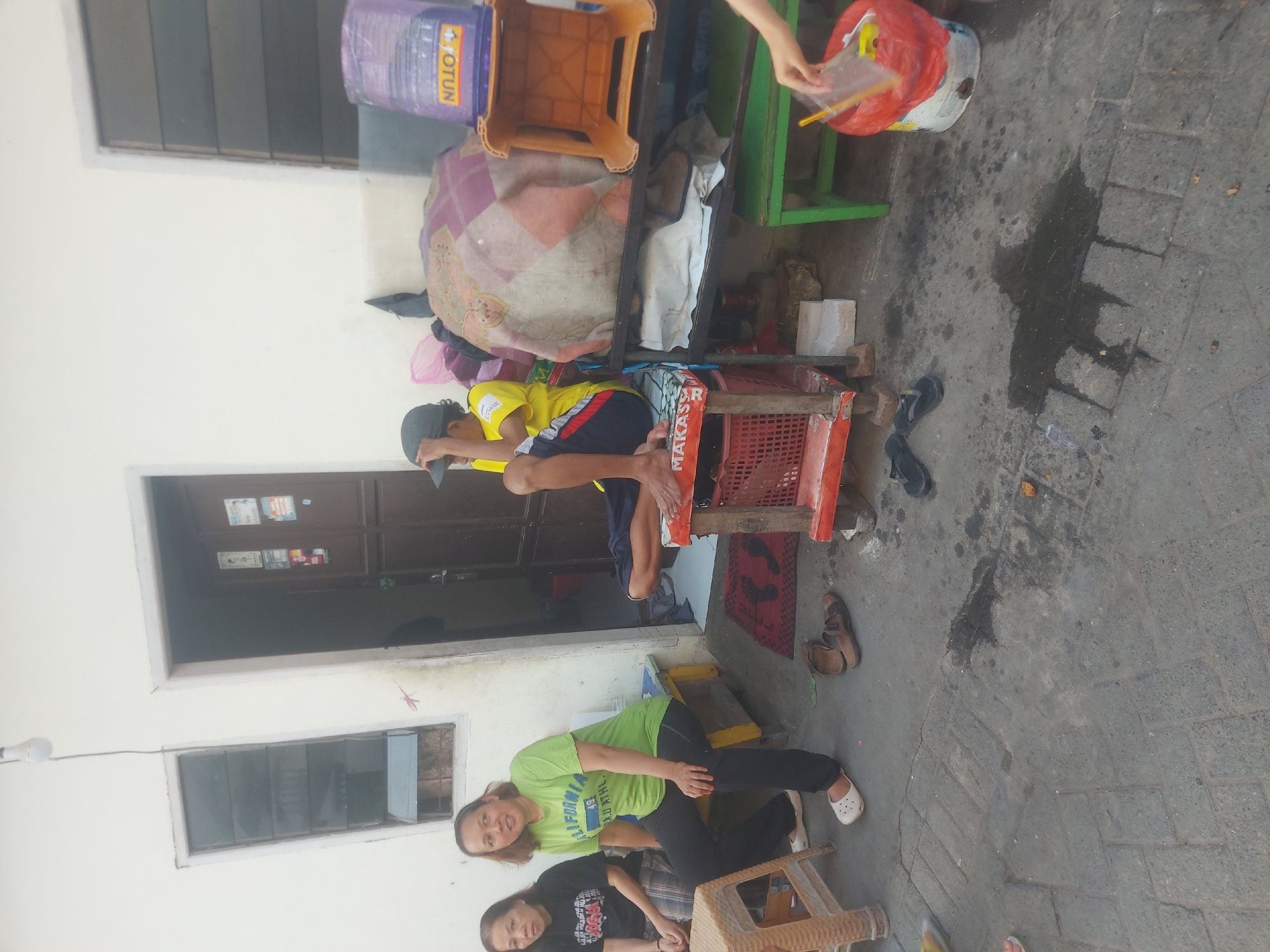 The image shows a street scene in Makassar, Indonesia, with several people gathered. In the foreground, the ground is paved with interlocking stones and shows some wet patches. There are several pairs of flip-flops on the ground.
To the left, two women are sitting on a yellow bench. The woman in the front is wearing a green t-shirt with "CALIFORNIA" and "ATHL." printed on it, black pants, and white clogs. The woman behind her is wearing a black t-shirt. A wicker stool is next to them.
In the middle of the image, a young person wearing a yellow t-shirt and dark shorts is sitting on a red crate. They are wearing a dark baseball cap.
Behind them, there is a shelf unit with various items, including a large purple water jug with "OTUN" printed on it and an orange plastic stool. A colorful patterned blanket is also visible.
To the right, a red bucket is placed on the ground, and a hand is reaching into it, holding a plastic bag with some items inside, possibly a toy car. A green bench is visible behind the red bucket.
The background features the side of a building with a window and a closed door. The overall impression is of a casual gathering in an outdoor urban setting.