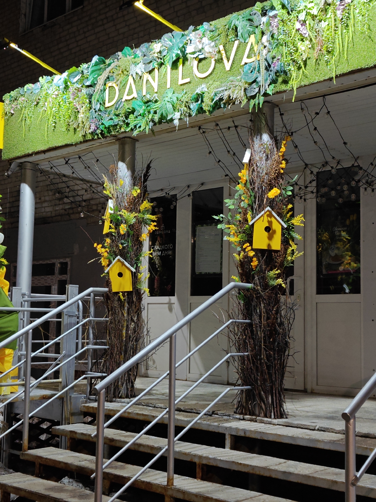 The image shows the entrance to "Danilova," possibly a shop or restaurant, located in the city of Chaikovsky, Russia. The entrance has metal stairs with silver railings leading to a covered porch. Two faux tree trunks adorned with yellow flowers and small yellow birdhouses flank the entrance. A sign above the entrance reads "DANILOVA" in gold lettering against a backdrop of lush artificial greenery. Fairy lights are strung across the porch roof. The building behind the entrance appears to be brick. The scene is lit, likely at night.