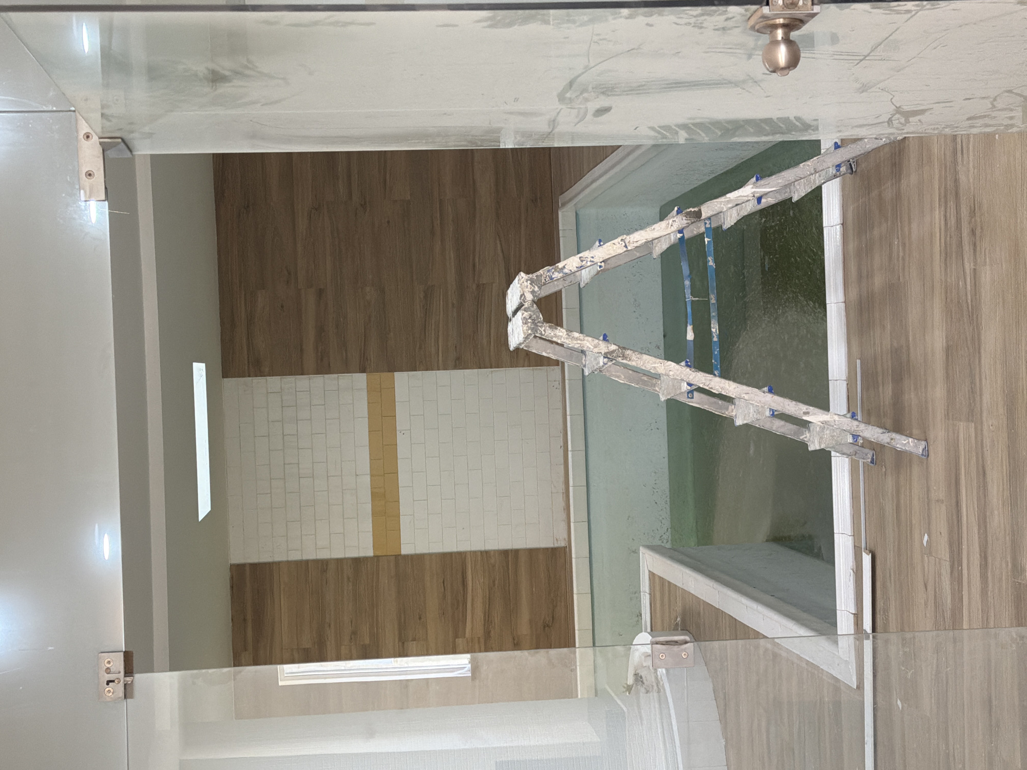 The image depicts an indoor swimming pool area under construction, likely a mikvah, situated in Gondomar, Portugal. A silver aluminum stepladder, showing signs of paint splatters, stands open and centered within the pool's enclosure. The pool itself is filled with greenish water and appears to have a concrete basin. The surrounding walls are finished with a combination of wood-effect paneling and white subway tiles, accented by a horizontal band of yellow tiles. The floor is covered in wood-look laminate flooring. A glass partition with metal hinges is visible on the left, and a doorframe with a brass doorknob is on the right, suggesting a completed section of the building. The ceiling features recessed lighting. There are no people present in the image.