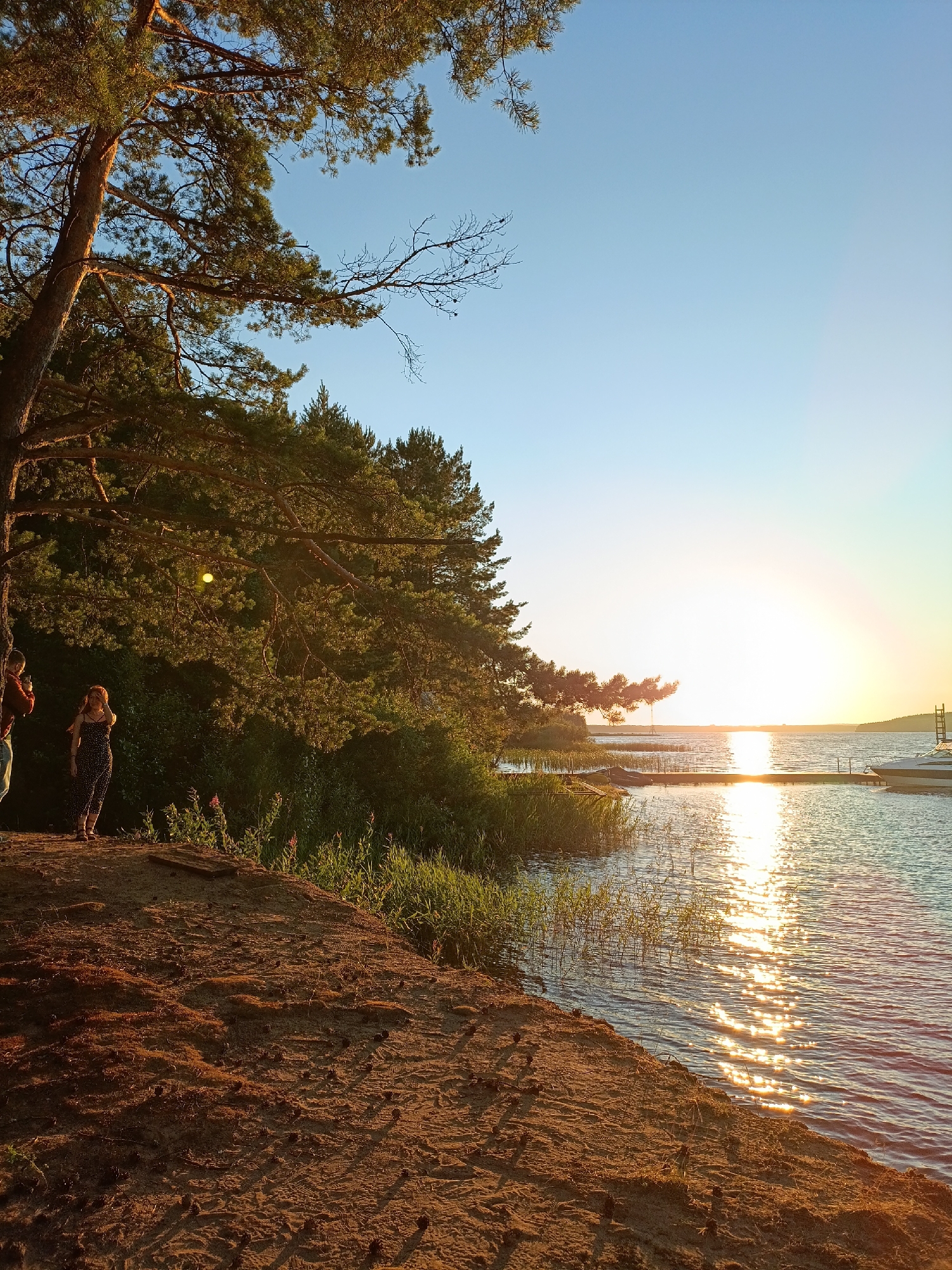The image depicts a scenic sunset view on the Kama River in Chaykovsky, Russia. In the foreground, a sandy embankment is covered with pine cones and sparse dry grass. Two individuals are visible on the embankment; one person, wearing a dark patterned dress, stands facing the water, while another person, partially obscured, is to their left. Lush green vegetation and pine trees line the embankment, with sunlight filtering through the branches. The river stretches out to the horizon, reflecting the golden hues of the setting sun, creating a shimmering effect on the water's surface. A wooden dock with a boat is situated on the right side of the river. The sky is a clear, pale blue, transitioning to a brighter yellow near the horizon where the sun is setting.