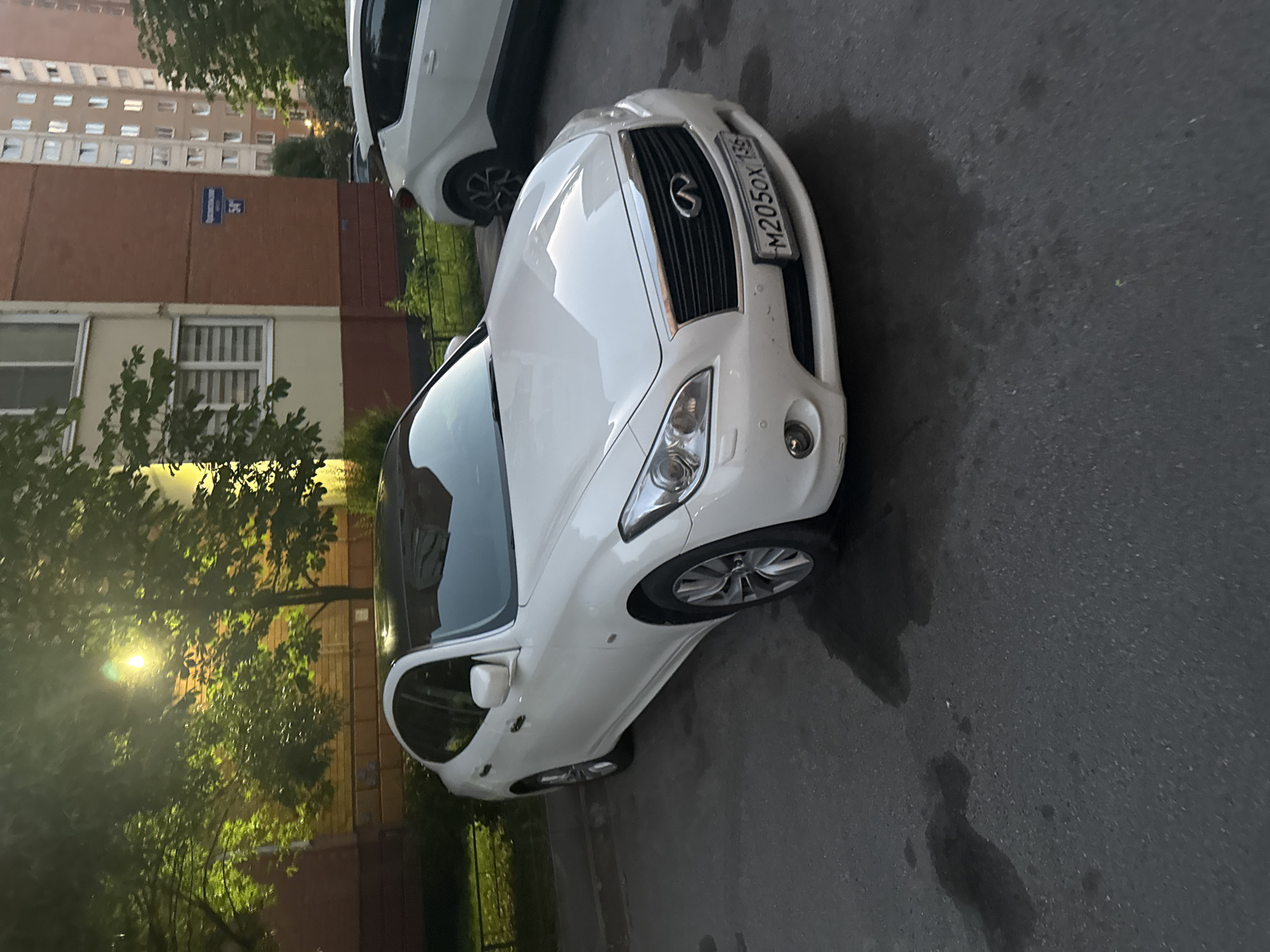 This media file captures a white Infiniti sedan parked on a wet asphalt street in Saint Petersburg, Russia. The car is viewed from a low angle, emphasizing its front end and the driver's side. The license plate is clearly visible and reads "M205OX 159".
The scene suggests it is late evening or early morning, as indicated by the warm glow of streetlights filtering through the lush green trees lining the street. The wet ground indicates recent rain or a damp environment. In the background, a brick building with multiple windows and a blue street sign is visible, adding context to the urban setting. The overall mood is quiet and still, with the parked car as the primary subject. There are no people visible in the frame.