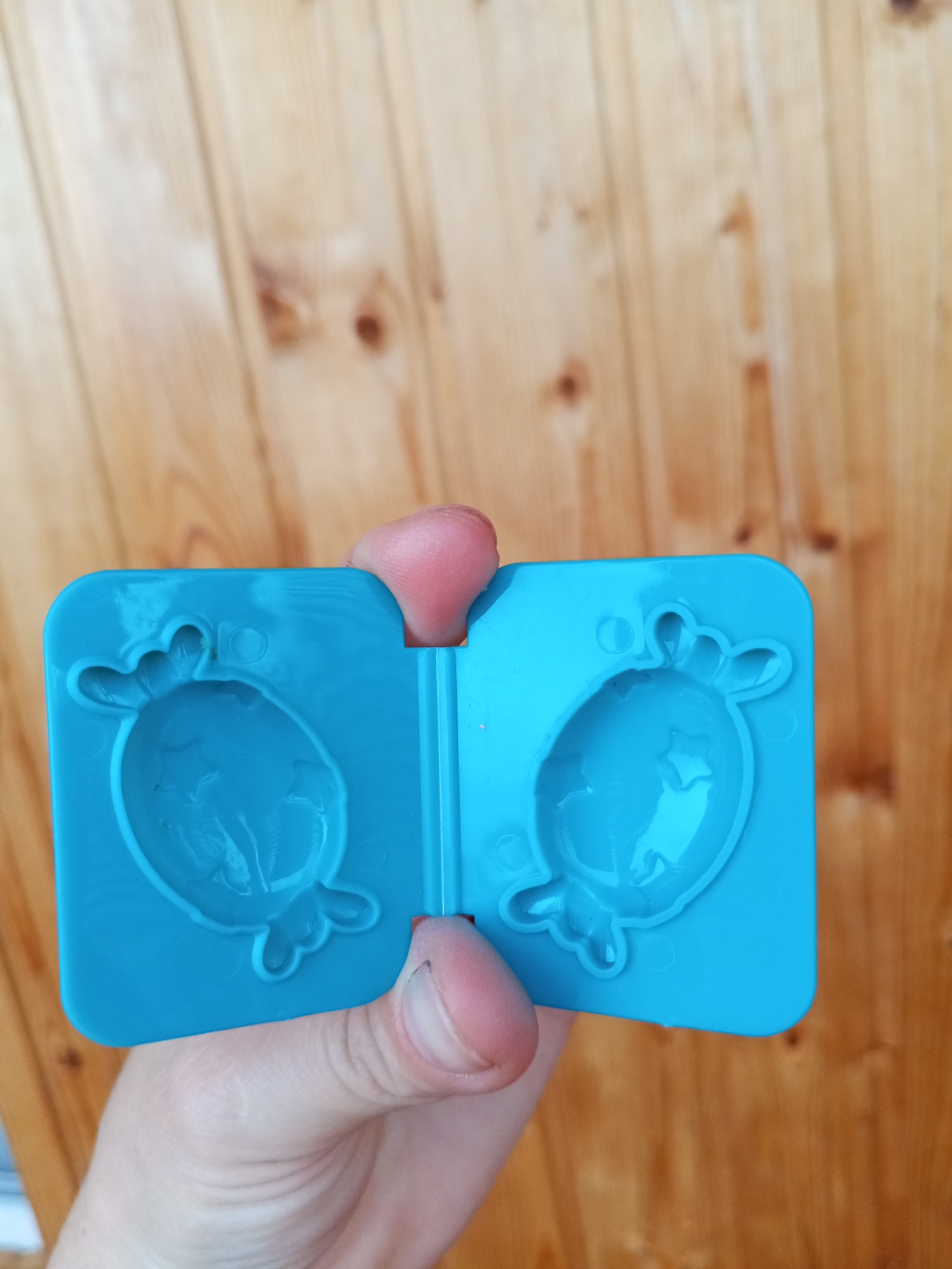 In Chistopol, Russia, a pair of bright blue silicone molds are held open by a person's hands against a background of light brown wooden planks. Each mold is shaped like a whimsical creature with a round body and four rounded protrusions at the top. The left mold shows a subtle indentation within its cavity, suggesting a detail or face. The person's thumbs are visible, pressing the edges of the molds apart, their skin showing a light tone and faint lines. The wooden planks are oriented vertically, with visible grain patterns and knot details. The lighting appears to be natural daylight, casting soft shadows.