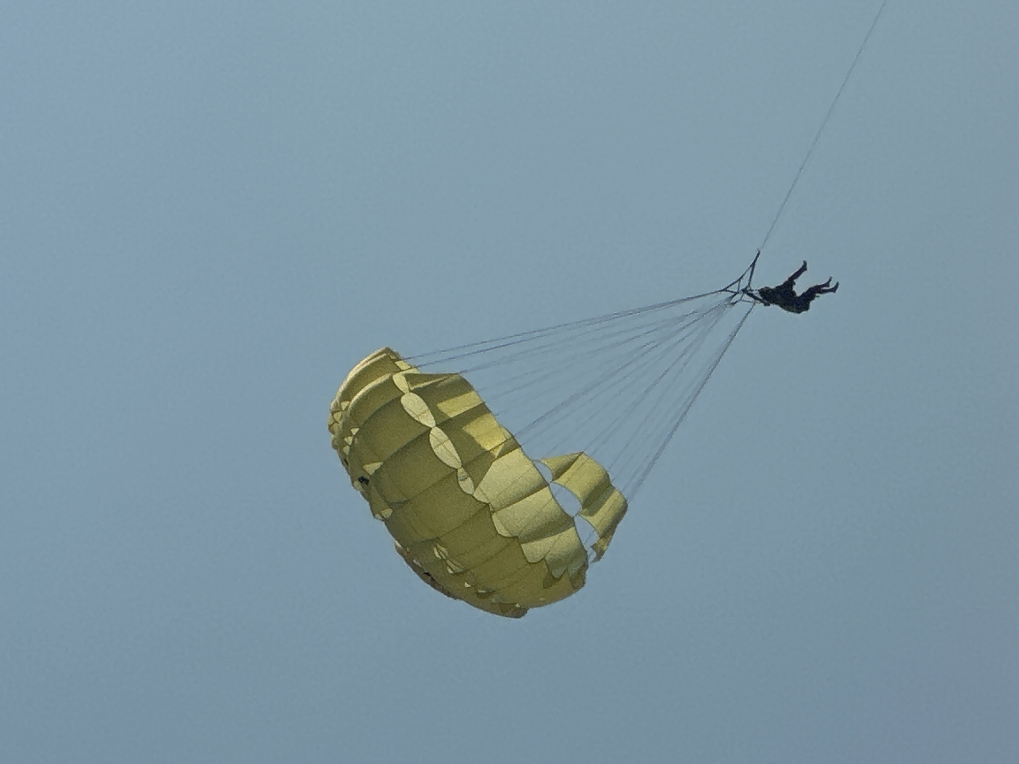 A person is parasailing in the sky above Boreti, Montenegro. The parasail is yellow and inflated, with the person hanging below attached by lines. The sky is a clear blue.