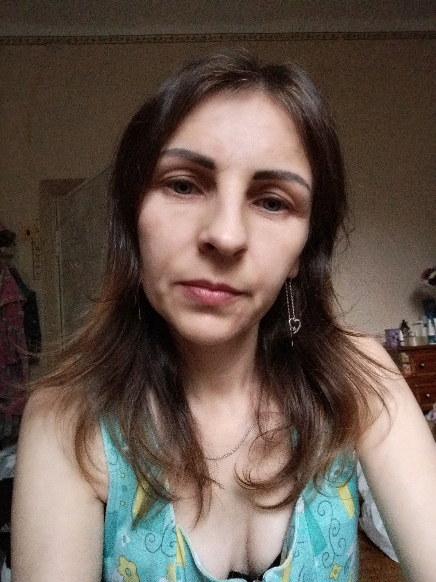 A woman is pictured from the chest up, looking directly at the camera. She has long, dark brown hair that is parted in the middle. She is wearing a light blue top with a floral pattern. She has a delicate silver necklace on. She is also wearing a long, dangling earring in her left ear that features a heart-shaped charm. The background is dimly lit and appears to be a room with furniture and clothing. There is no indication of the city of Vinnytsia, Ukraine, or any specific location.