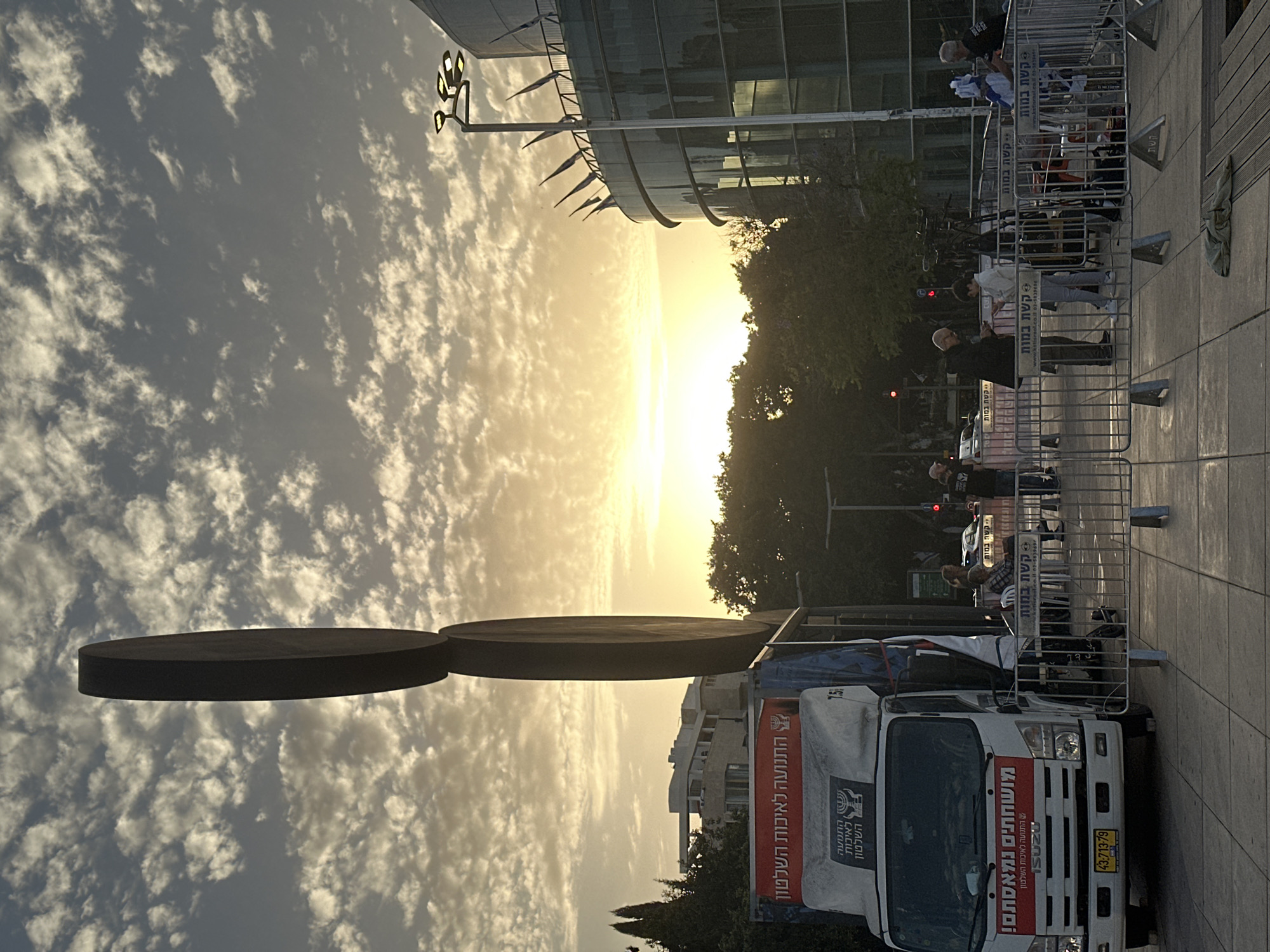Here is a detailed description of the image:
**General Scene:**
The image is taken at a street level, outdoors, likely during sunset or sunrise given the sky's golden hue.
**Foreground:**
- In the lower part, there's a white Isuzu truck with Hebrew lettering on its side and back. Text on the truck includes slogans like "מושחתים נמאסתם! " (Corrupt, we're tired of you!) and mentions "התנועה לאיכות השלטון" (The Movement for Quality Government). The license plate is visible.
- A two-disk-shaped piece of art that rises into the air is also visible.
- Barriers are set up to prevent people from passing.
- People are visible behind the barriers.
**Middle Ground:**
- A row of barriers with people behind them, potentially indicating a gathering or protest.
- Trees, possibly indicating a park or green area are present.
- Part of what appears to be a modern glass building is visible.
**Background:**
- A cloudy sky dominates the background, with the setting/rising sun creating a bright, golden glow near the horizon.
- Distant buildings are visible, suggesting an urban area.
**People:**
- Several individuals are visible behind the barriers, some appearing to be engaging in conversation or observing the scene.
**Objects:**
- Barriers (likely metal).
- A large white truck.
- Possible art in the form of a disk.
- Street lights.
**Location (Probable):**
Given the Hebrew text and context of the truck's message, this image is most likely taken in Israel. While not explicitly stated, the context of the truck's message and design could suggest this location. Based on the style of architecture and other details, it's probable that this is in Tel Aviv.