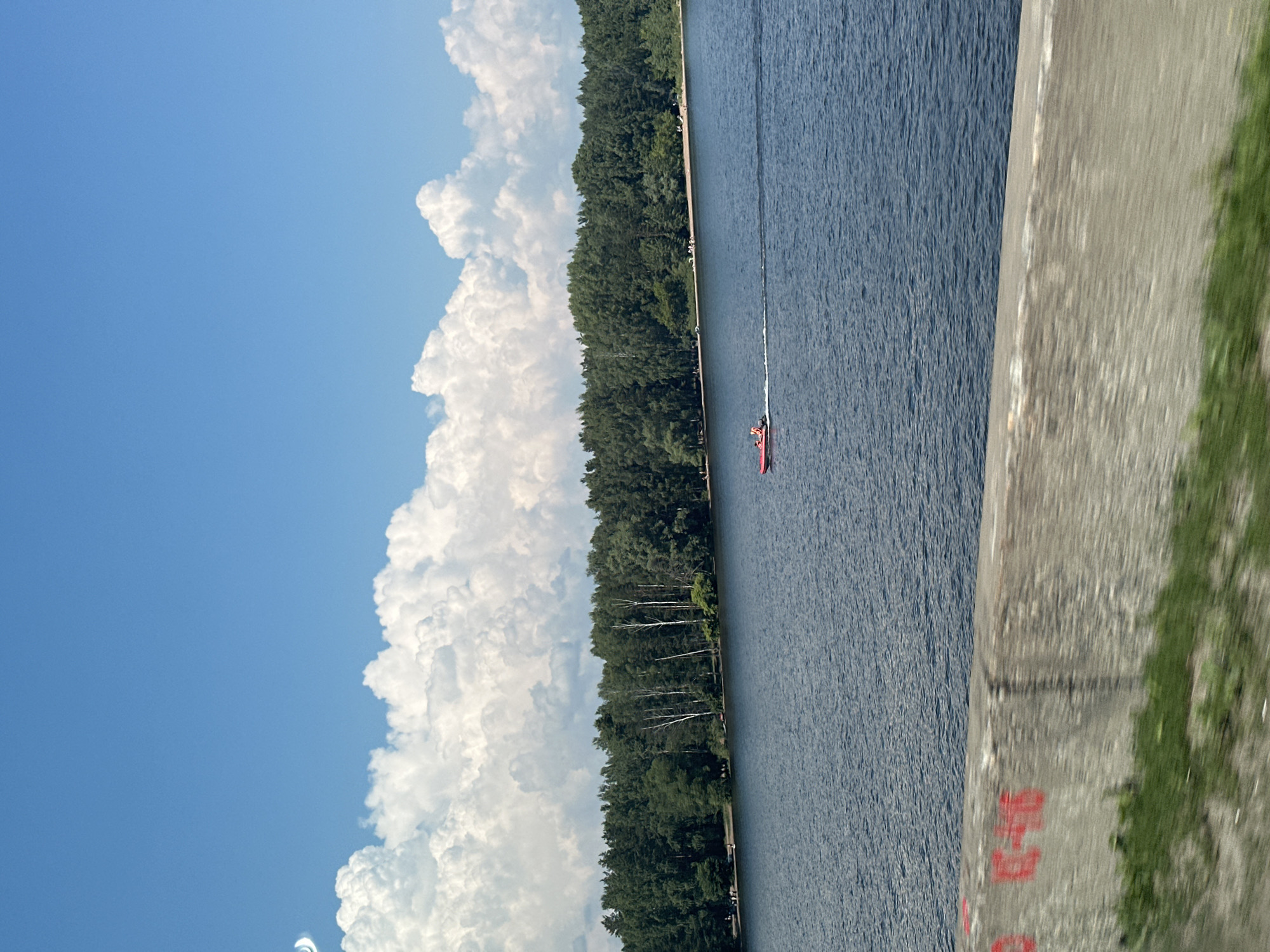 A wide-angle, eye-level shot depicts a serene lake scene under a bright blue sky dotted with fluffy white clouds. The water is a deep blue, with gentle ripples reflecting the light. On the far side of the lake, a dense line of green trees forms a natural border, with some taller birch trees interspersed among them. Along the shoreline, small groups of people are visible, suggesting a park or recreational area. A red speedboat, with two occupants, is actively moving across the water, leaving a white wake behind it. In the foreground, a concrete barrier with red graffiti marks runs horizontally across the bottom of the frame, with blurry green grass visible below it, implying the photo was taken from a moving vehicle or elevated vantage point.