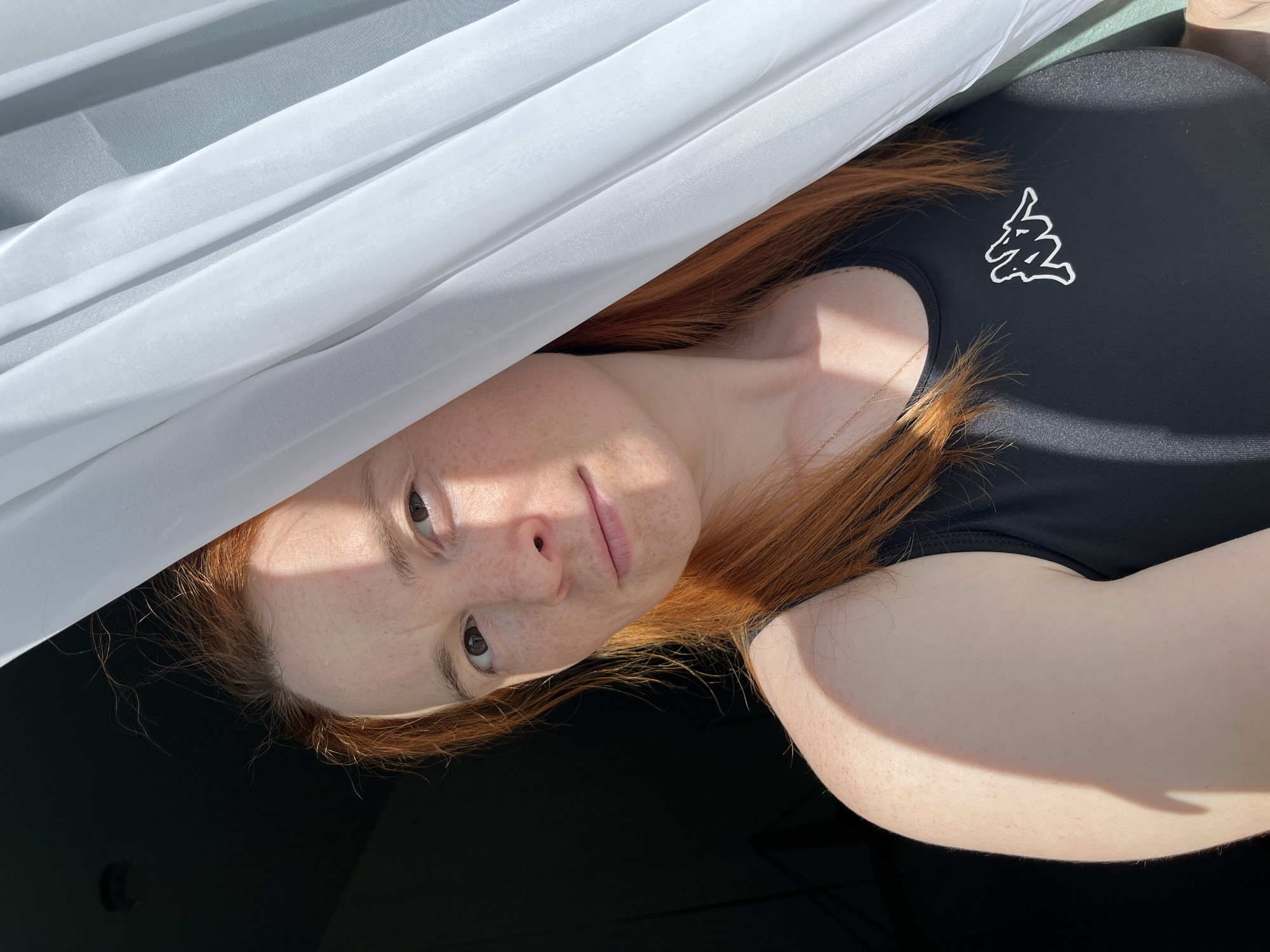 This is a close-up, horizontal, portrait-style photograph of a young woman with long, reddish-brown hair. She is lying down and looking towards the camera, with a portion of her face, particularly her eyes and forehead, illuminated by bright sunlight, casting distinct shadows. She has fair skin with freckles across her nose and cheeks. A delicate gold chain necklace is visible on her décolletage.
She is wearing a black, short-sleeved top with a small, white, stylized logo on the left side of her chest, resembling a leaping animal or a dynamic abstract shape. The background is divided into two distinct areas: the upper portion features draped white and light grey fabric, possibly a curtain or sheer material, catching the light and creating soft folds. The lower part of the image is a dark, solid surface, likely a table or floor.
The lighting suggests it is daytime, possibly midday, with strong sunlight. The overall mood is relaxed and intimate, with the subject appearing calm and direct in her gaze. There is no visible text in the image. The context provided indicates this scene is in Chaikovsky, Russia.