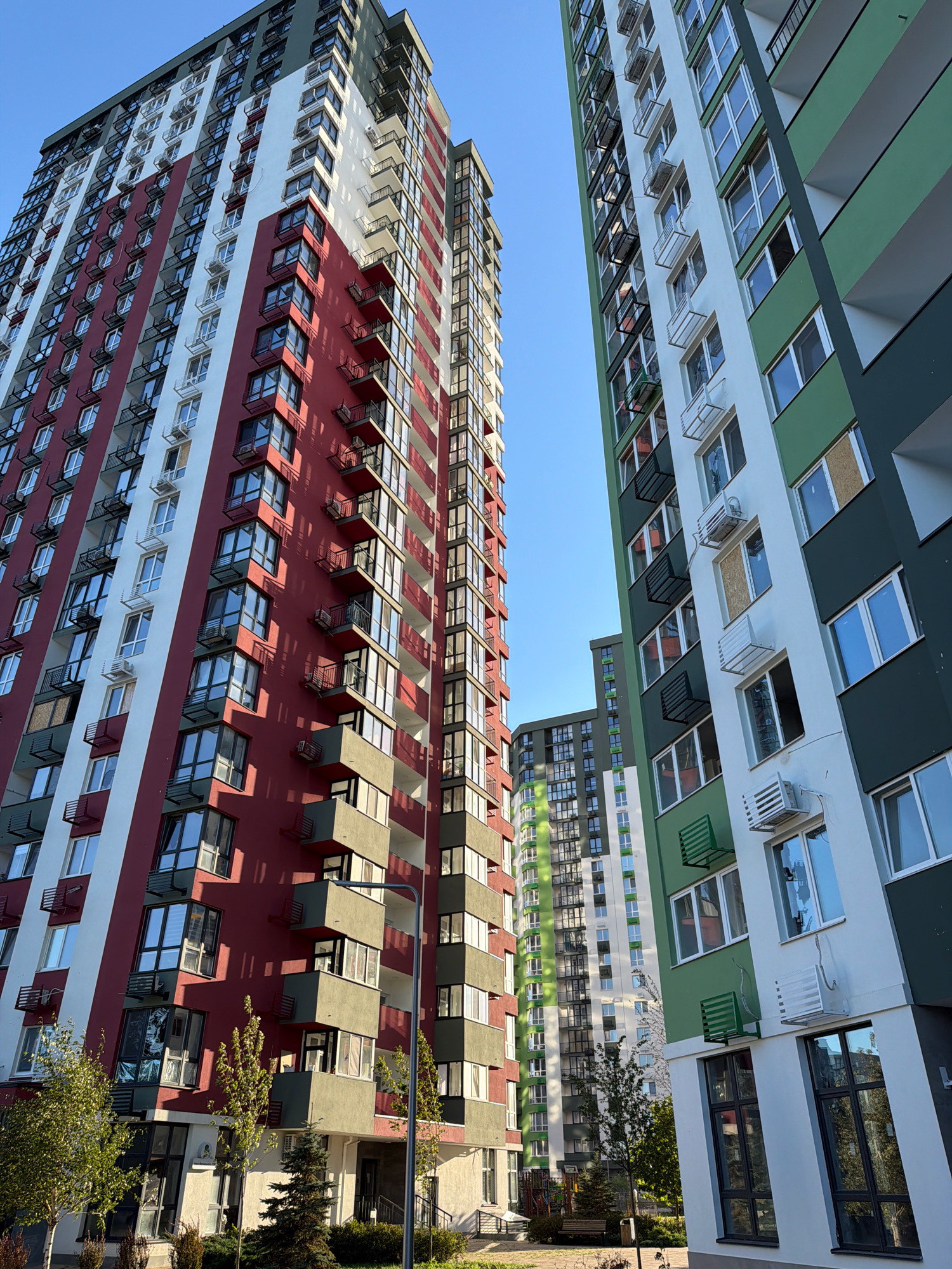 The image shows a modern residential complex in Kyiv, Ukraine, featuring several high-rise buildings with distinct architectural styles and color palettes. The buildings are tall, multi-story structures with numerous windows and balconies. One building on the left is primarily red and white, with a section of gray on the lower floors. Another building on the right is white and green, with a dark green facade. A third building, in the background, appears to be a mix of white and green. The sky is clear and blue. In the foreground, there are trees, bushes, and some landscaping. A lamppost is visible near the red and white building. The scene appears to be a sunny day.