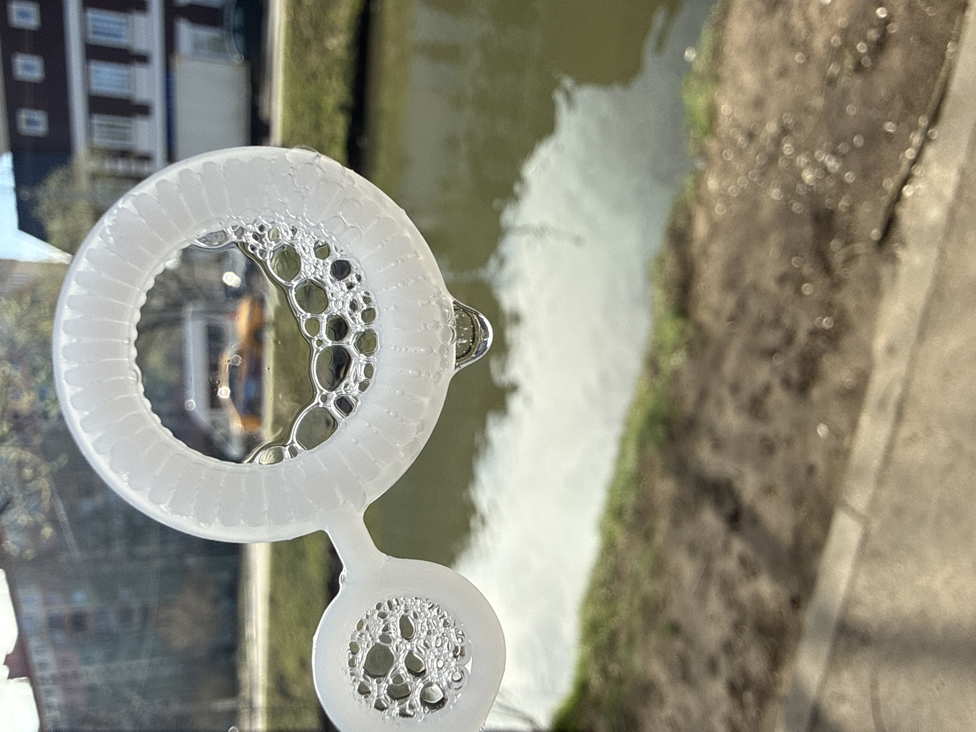 A plastic bubble wand with soapy water films forming bubbles is in the foreground. Behind the wand, a canal is visible, reflecting the sky and possibly a building. On the right side of the frame, there is a sidewalk and vegetation along the canal. A building with multiple windows is partially visible in the background. The location is plausibly Dmitrov, Russia, given the architectural style of the building and the presence of canals in the area. No people are directly visible.