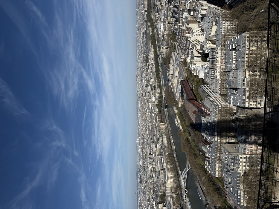 This high-angle shot captures a vast panorama of Paris, France, from an elevated position on the Eiffel Tower, looking predominantly northeast. The foreground features a dark metal railing from the observation deck and the prominent, elongated shadow of the Eiffel Tower itself, stretching diagonally across a cluster of residential and commercial buildings directly below. The shadow obscures parts of the rooftops and facades, indicating strong, direct sunlight.
The middle ground is dominated by the Seine River, flowing from right to left, with several barges visible on its surface. Multiple bridges span the river; one distinctly features white arches, likely the Passerelle Debilly, and another, further downriver, appears to be the Pont de Bir-Hakeim. The riverbanks are lined with trees, some still bare of leaves, and densely packed Haussmannian buildings with their characteristic grey rooftops. On the left bank, notable structures include the Palais de Tokyo and the Musée Galliera.
The background extends into a sprawling urban landscape of Paris, characterized by an endless expanse of uniformly colored buildings under a clear, bright blue sky. Scattered white, wispy cirrus clouds and faint contrails are visible in the upper atmosphere. Distant landmarks include the Montparnasse Tower appearing as a light-colored rectangular skyscraper towards the right horizon, and the towers of La Défense business district visible towards the left-center horizon. No individual people or explicit interactions are discernible due to the scale of the view. The scene is illuminated by strong daylight, casting sharp shadows.