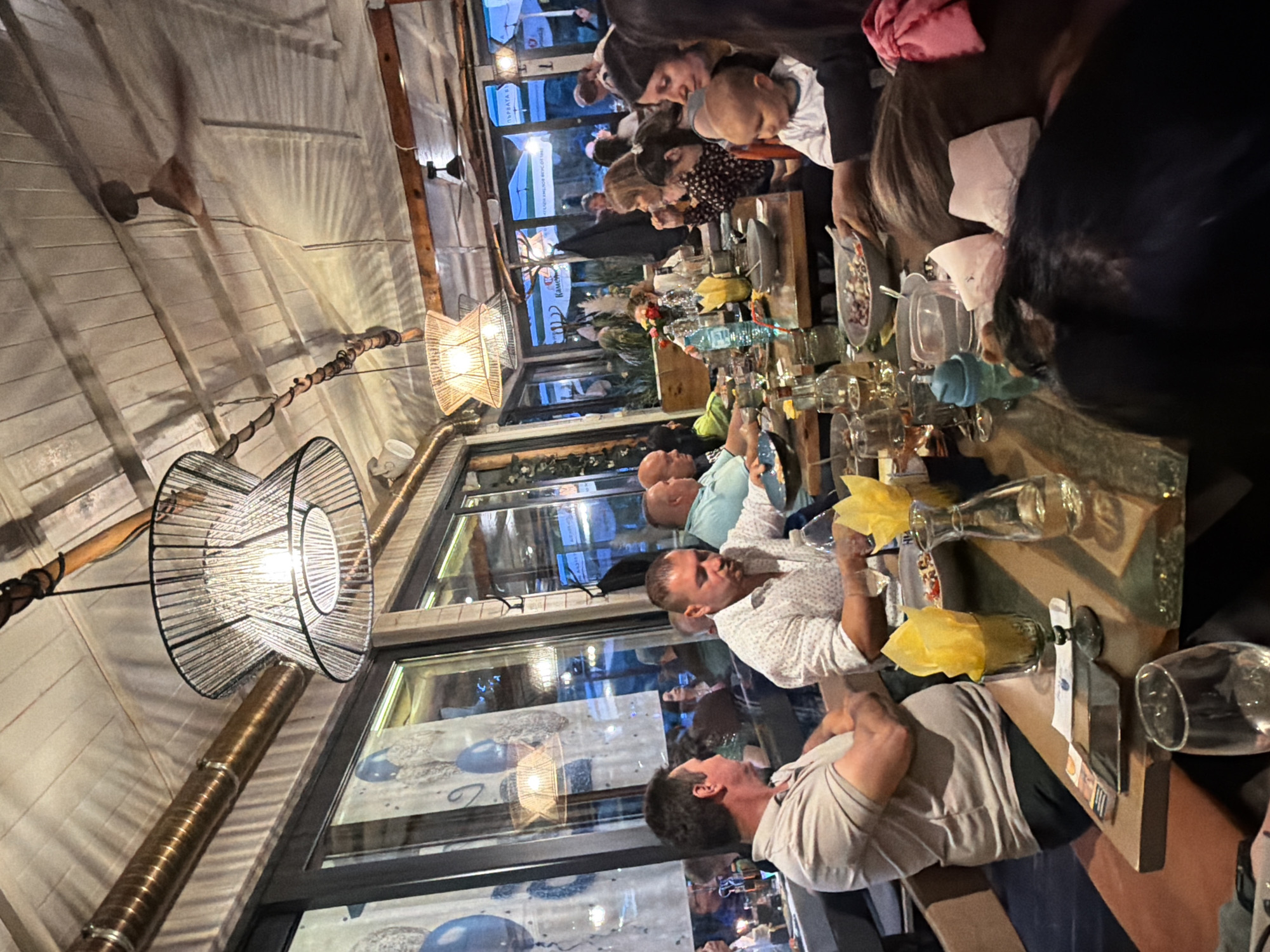 The image depicts a lively gathering of people seated at a long, wooden table inside a restaurant or bar. The location is suggested to be Burgas, Bulgaria, potentially a waterfront venue, given the blurred view of the outdoors visible through the large windows behind the diners. The windows are decorated with marine-themed art, featuring jellyfish.
The lighting is primarily from overhead light fixtures, including a distinctive, large, spherical wireframe chandelier. Some of the diners are engaged in conversation, with individuals holding glasses and what appear to be napkins. The table is set with glassware, including wine glasses and bottles of water.