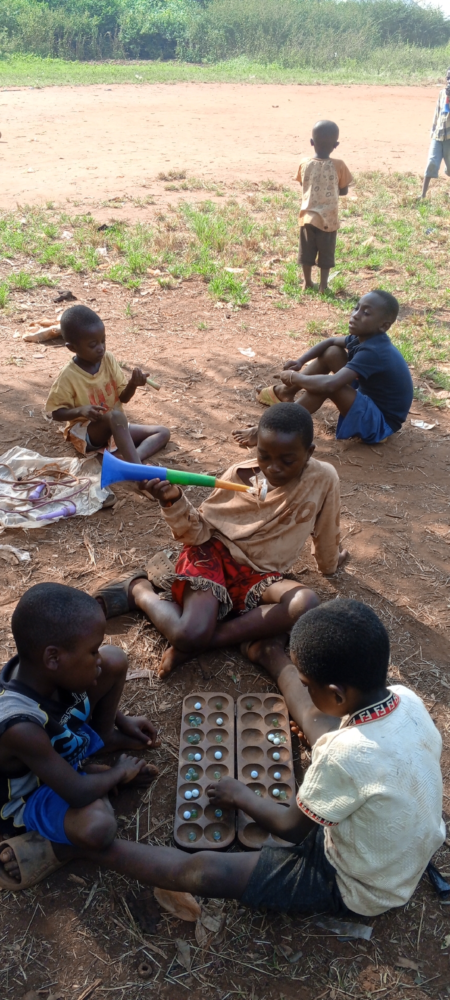 In Ngalula, Democratic Republic of the Congo, six African children are situated on reddish-brown soil scattered with small patches of green grass, twigs, and leaves. The children are positioned in a partial circle. Two are engaged in playing a traditional board game, likely Mancala or a similar variant, on a wooden board with small divots filled with marbles. One child holds a blue and green vuvuzela-like horn. Another child sits nearby with a similar, smaller, unicolored instrument. A length of braided cord is near the vuvuzela. One child is sitting apart, observing the others. Another child is standing alone in the background. The backdrop features sparse shrubbery and a dirt road.