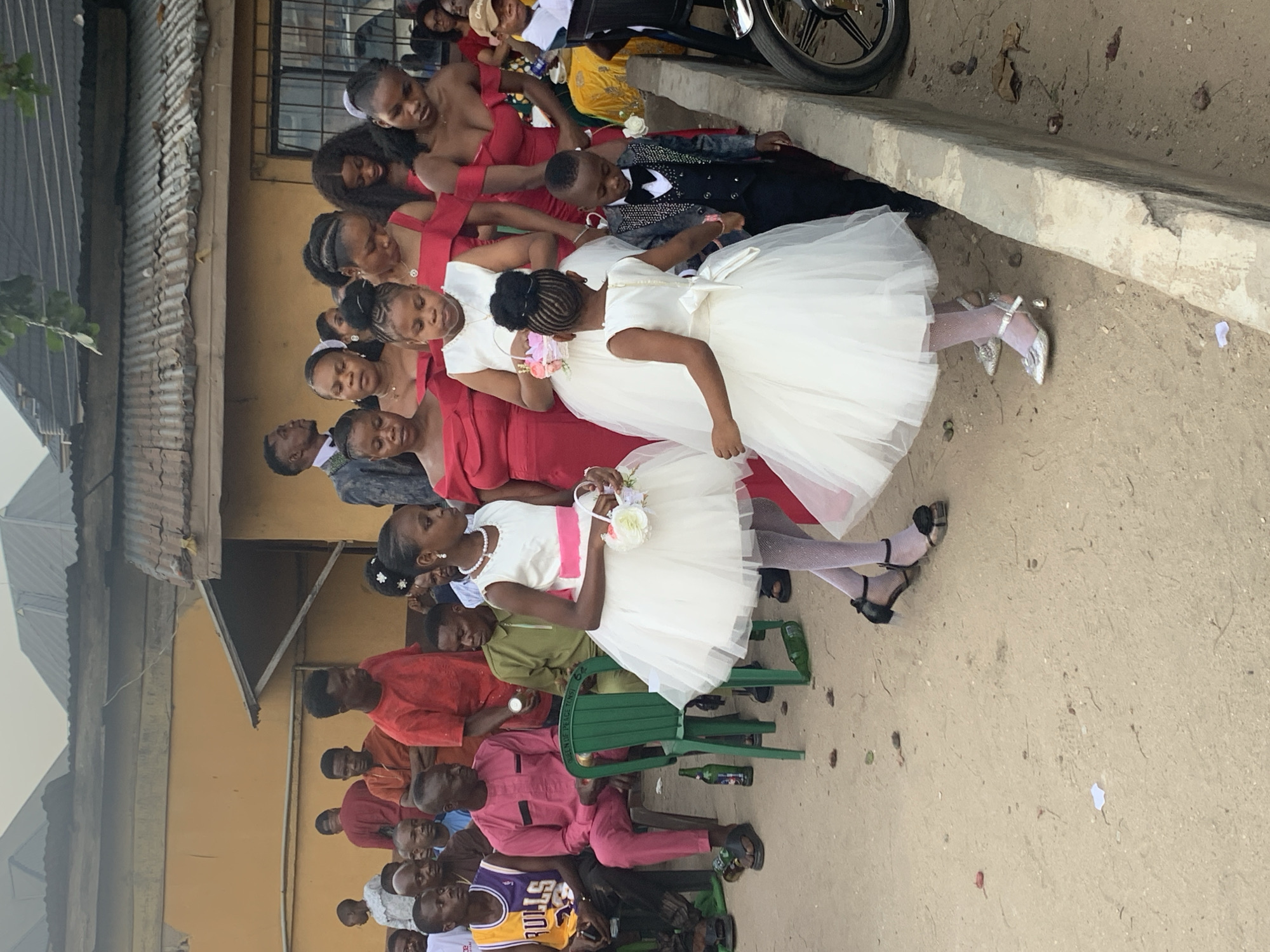 The image captures an outdoor social gathering in Umuode, Nigeria. In the immediate foreground, three young girls in white, layered dresses are visible. The leftmost girl wears a white dress with a pink sash, white patterned tights, and black flat shoes, holding a small white floral basket. The central girl, facing left, wears a white dress with a white sash, white patterned tights, and silver low-heeled shoes, also holding a small white floral basket. She is holding the right hand of a young boy to her right. This boy wears a dark grey sequined suit jacket, a black vest, black trousers, and a black bow tie.
Behind these children, multiple adult women are present, uniformly dressed in off-the-shoulder red formal gowns. Further into the midground and background, numerous men are gathered. Some men are seated on green plastic chairs; one in a pink long-sleeved tunic and matching trousers, and another in an olive-green long-sleeved tunic. Another man in the midground wears a purple and yellow "BULLS 23" basketball jersey. Other men stand or observe in the background, one visible in a red t-shirt and another in a grey patterned suit jacket.
The ground surface is unpaved, consisting of light brown dirt or sand, with scattered leaves and small pieces of debris. Empty green glass bottles are visible on the ground near the plastic chairs. A low concrete curb or barrier runs along the right side of the frame, with the front wheel of a motorcycle partially visible beyond it. The background consists of single-story buildings with mustard-yellow painted walls and weathered corrugated metal roofs. Sparse green foliage is visible overhead in the upper right. The lighting suggests daytime.