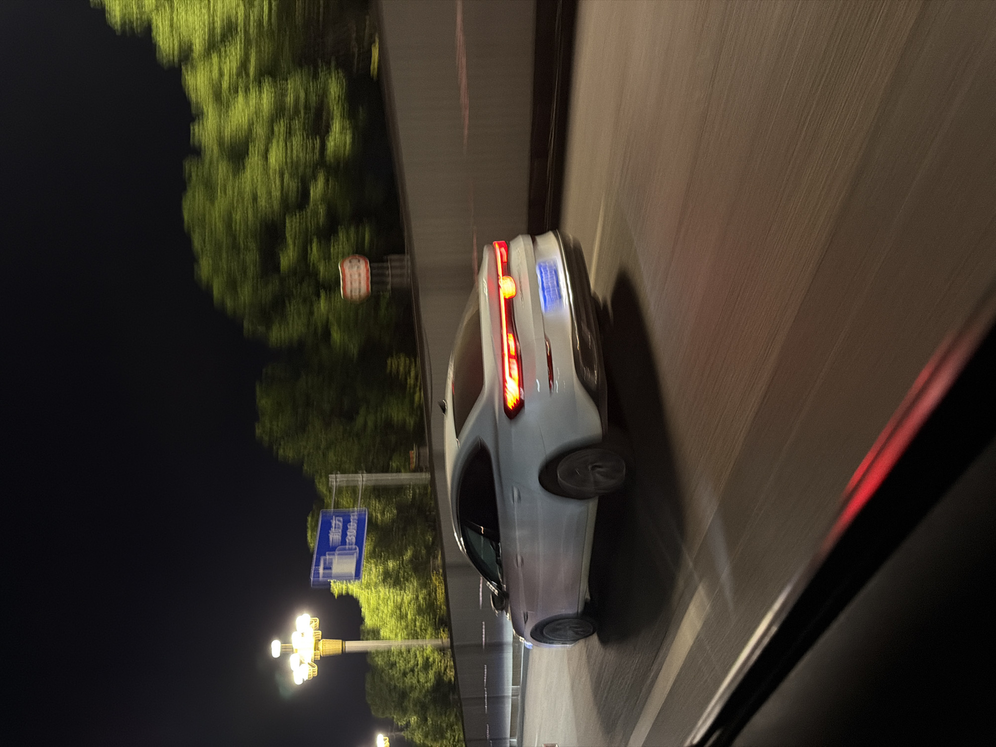 The image depicts a white sedan driving on a highway at night, presumably in Jiangjin, China, given the presence of a Chinese road sign. The car is in motion, as indicated by the motion blur on the trees and road. The car's brake lights are illuminated. A street lamp illuminates the left side of the scene, and trees line the right side of the highway behind a retaining wall. The road appears to be multi-lane.