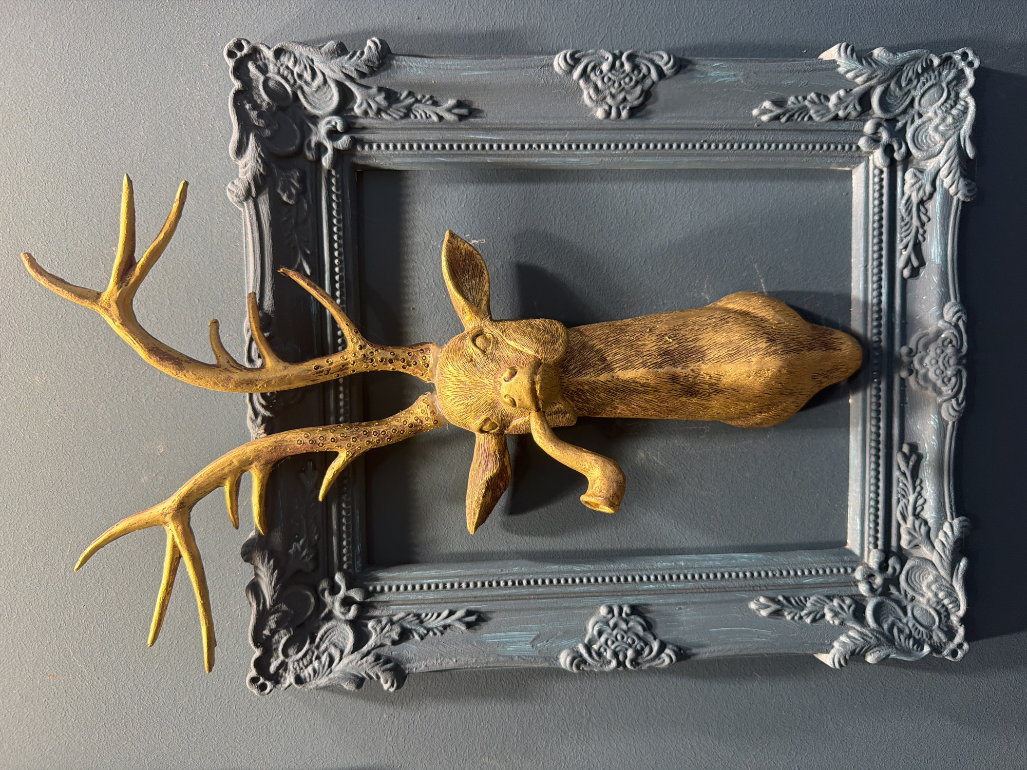The image displays a decorative golden deer head mounted on a teal-colored wall. The deer head features elaborate antlers and a pipe protruding from its mouth. It is positioned within an ornate, dark teal frame with intricate carvings. The wall has a textured finish. The image does not depict any specific location in the city of Lebedyan, Russia, nor does it show any people or actions.
