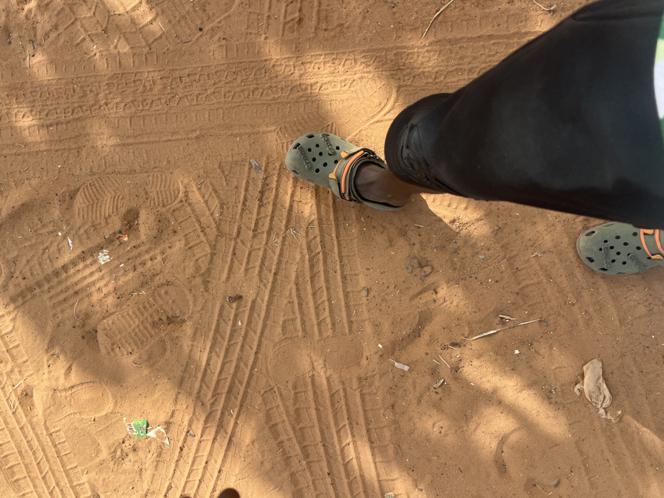 The image displays a ground-level perspective of an unpaved, sandy surface, likely a path, in Birnin Kebbi, Nigeria. A person's lower extremities are visible in the foreground, consisting of dark-colored trousers and two olive green Crocs-brand clogs with orange heel straps. The left foot is partially cropped at the bottom of the frame, while the right foot is more fully depicted. The reddish-brown sand substrate features numerous tire tracks from various vehicles, multiple human footprints (including distinct shoe prints and fainter barefoot impressions), and sparse natural debris consisting of dry plant matter. Scattered anthropogenic litter, including small white and green plastic fragments, is also present. Harsh shadows indicate direct overhead sunlight, characteristic of a daytime outdoor scene.