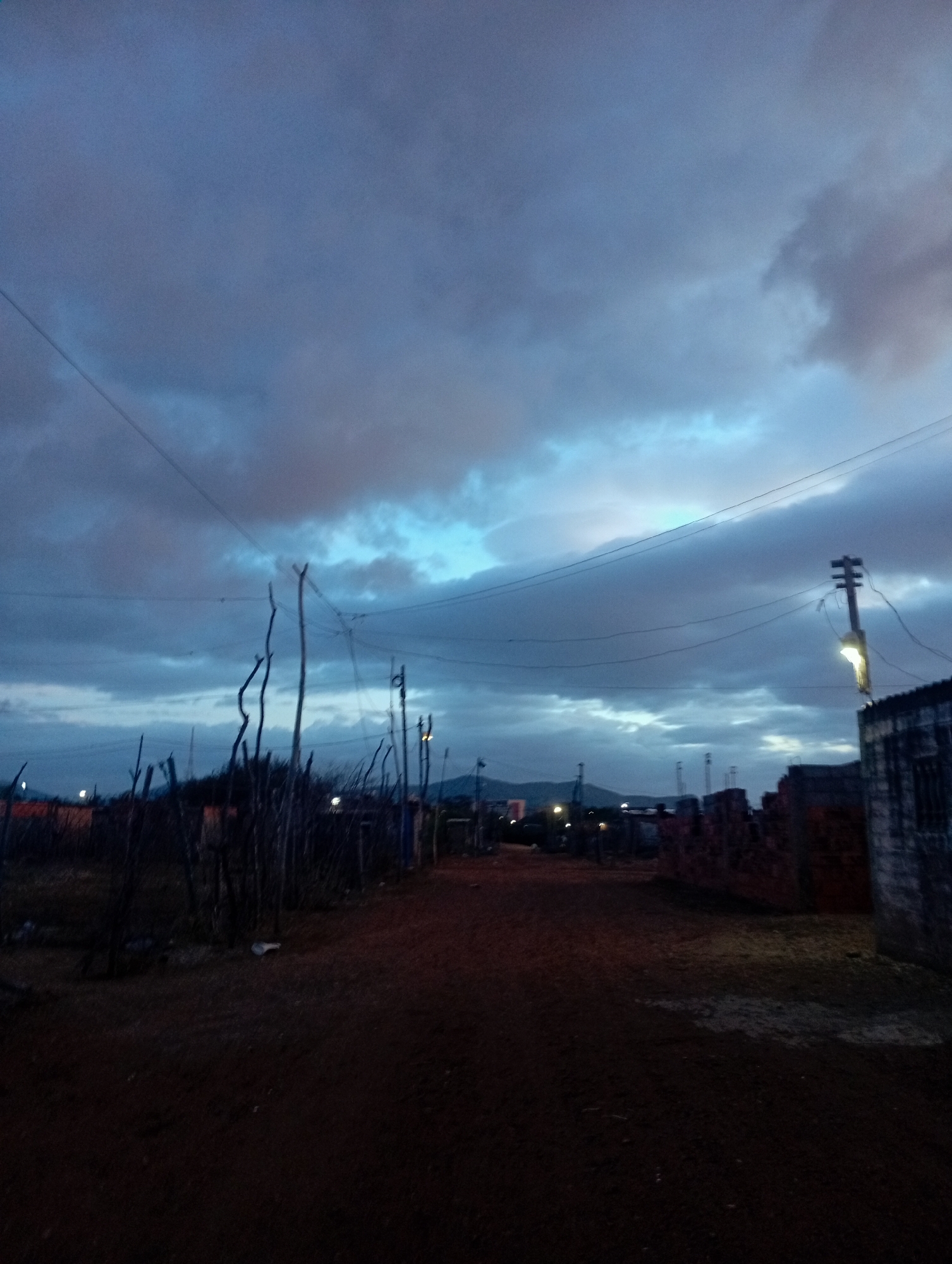 The image captures a dirt road in what appears to be a rural or semi-developed area, possibly at dusk or dawn. The sky is filled with dramatic, cloudy formations, with patches of lighter blue peeking through. Several utility poles and wires are visible, indicating the presence of infrastructure. On the sides of the road, there are sparse structures, including some made of wood or branches, and piles of bricks, suggesting construction or unfinished buildings. A single street light is illuminated on a pole to the right, casting a soft glow. The ground is a reddish-brown dirt, with some lighter patches. In the distance, a faint outline of hills or mountains can be seen. The overall atmosphere is somber and quiet, characteristic of a developing area.