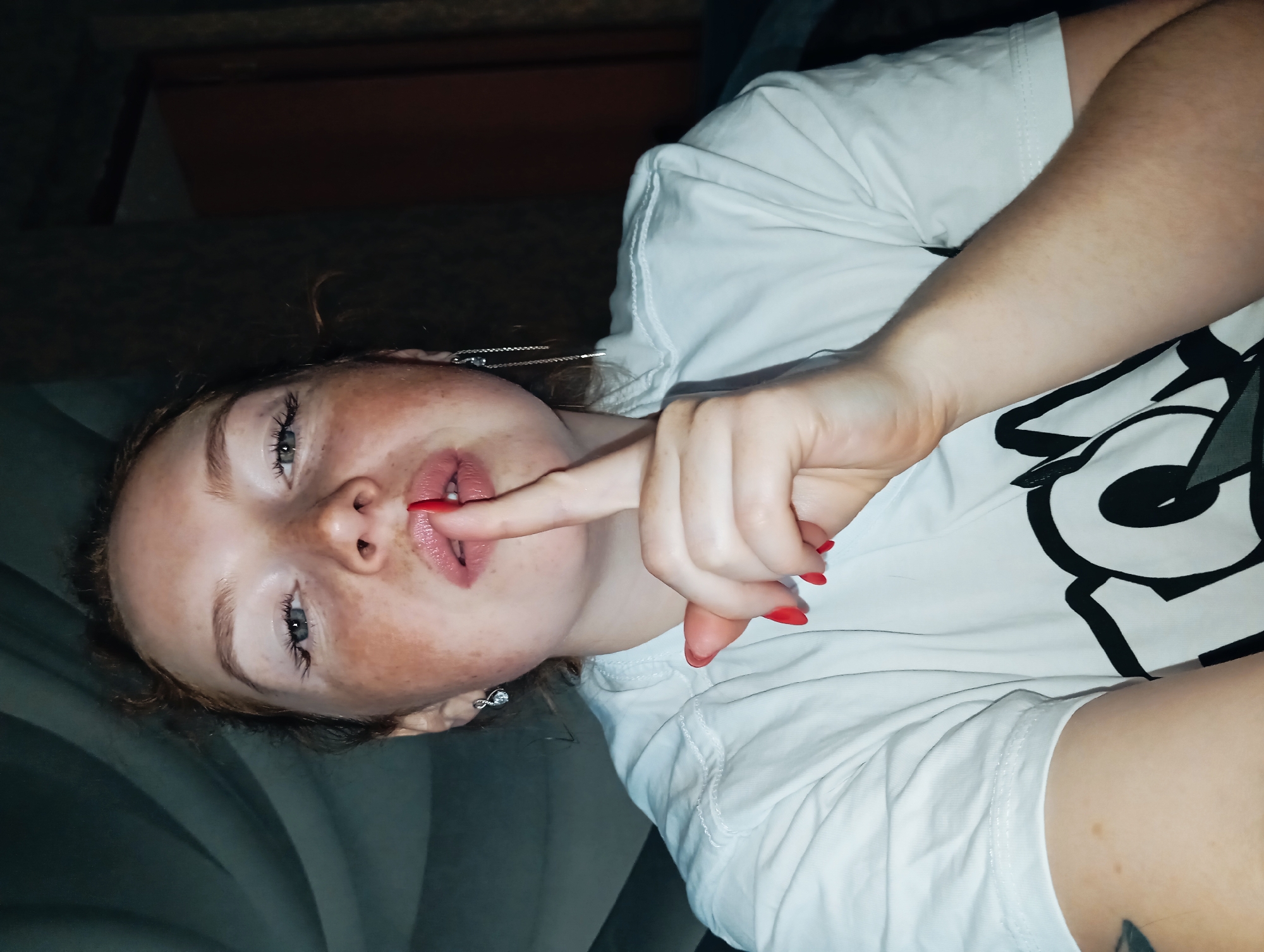 A pale-skinned person with freckles, wearing a white t-shirt with a black graphic design on the upper right, is lying on a gray upholstered surface. The person has red nail polish on their fingernails and is holding their index finger to their lips. The location is not discernible as Yurgamysh, Russia or any other location from the details provided in the image. The image appears to be taken indoors.