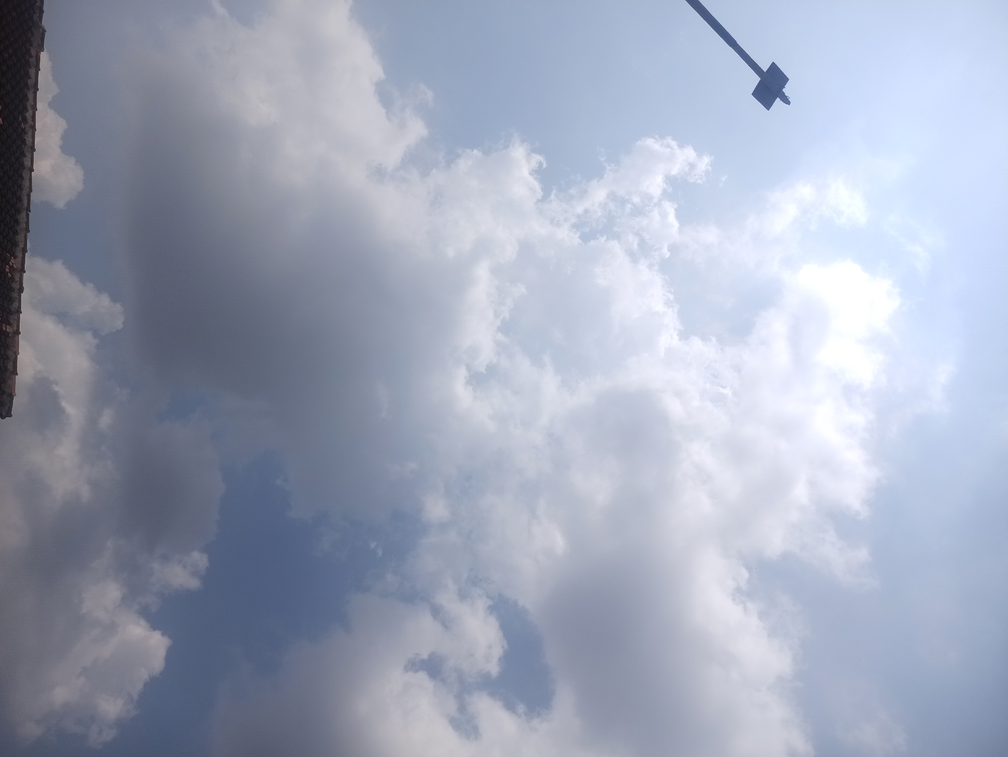 The image captures a sky filled with clouds, with the sun partially visible, creating a bright, washed-out area at the top left of the frame. A dark, vertical pole, likely a lamppost or a part of a structure, extends from the left edge of the image upwards into the sky. The bottom edge of the image shows a portion of a tiled roof, suggesting the scene is from ground level or a slightly elevated viewpoint. The clouds are varied in density, with some appearing wispy and others more substantial and shadowed, indicating a dynamic weather condition. The overall impression is that of an outdoor scene on a bright, cloudy day in Tuba, Indonesia, with a man-made element piercing the natural expanse of the sky.