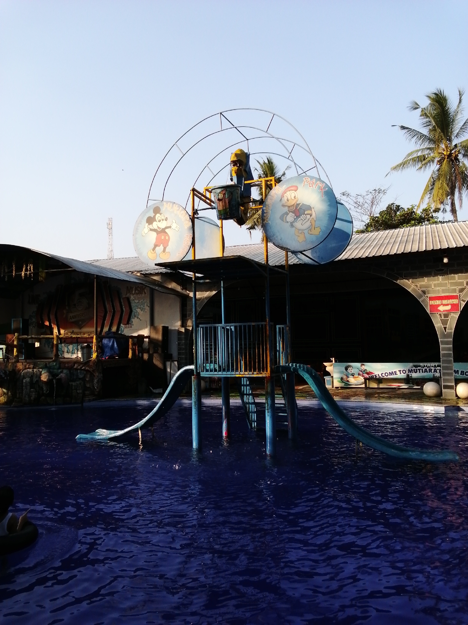 This image depicts a water park slide structure with Mickey Mouse and Donald Duck imagery, located in Kebonsari, Indonesia. The slide, made of blue and green plastic, is positioned over a deep blue pool of water. The structure is supported by blue poles, and a metal fence encloses the top. In the background, a building with a corrugated metal roof houses a stage with "MUSIK ANNIVERSARY" signage. Palm trees and a clear sky are visible above. The scene suggests a recreational environment, possibly during the day, with the pool water appearing calm and reflective.