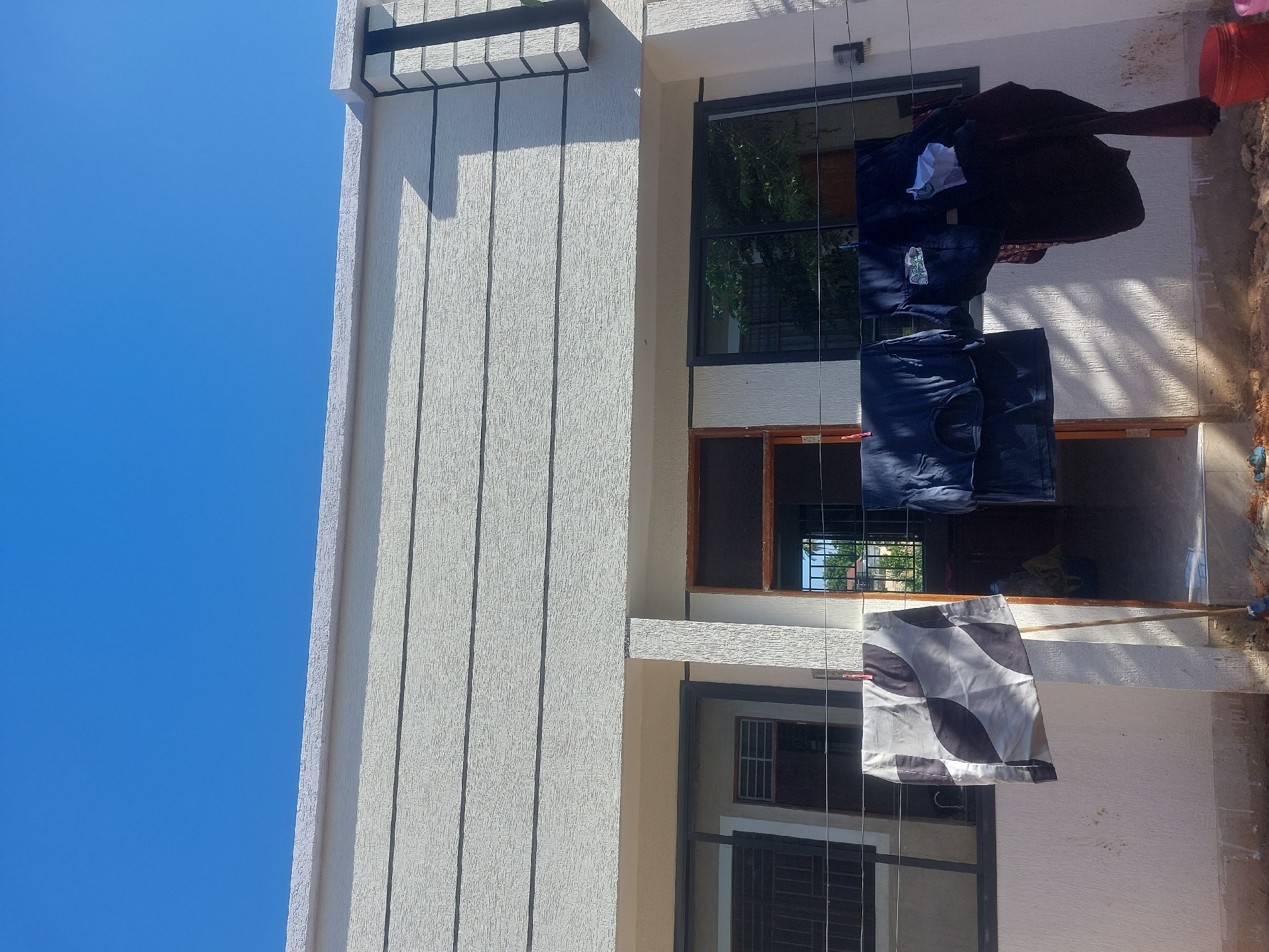 The image depicts a building under a clear blue sky. Clothes are hanging on a clothesline strung between the building's floors. The building has a textured exterior, with dark-framed windows. The location of the building is Dar es-Salaam, Tanzania.