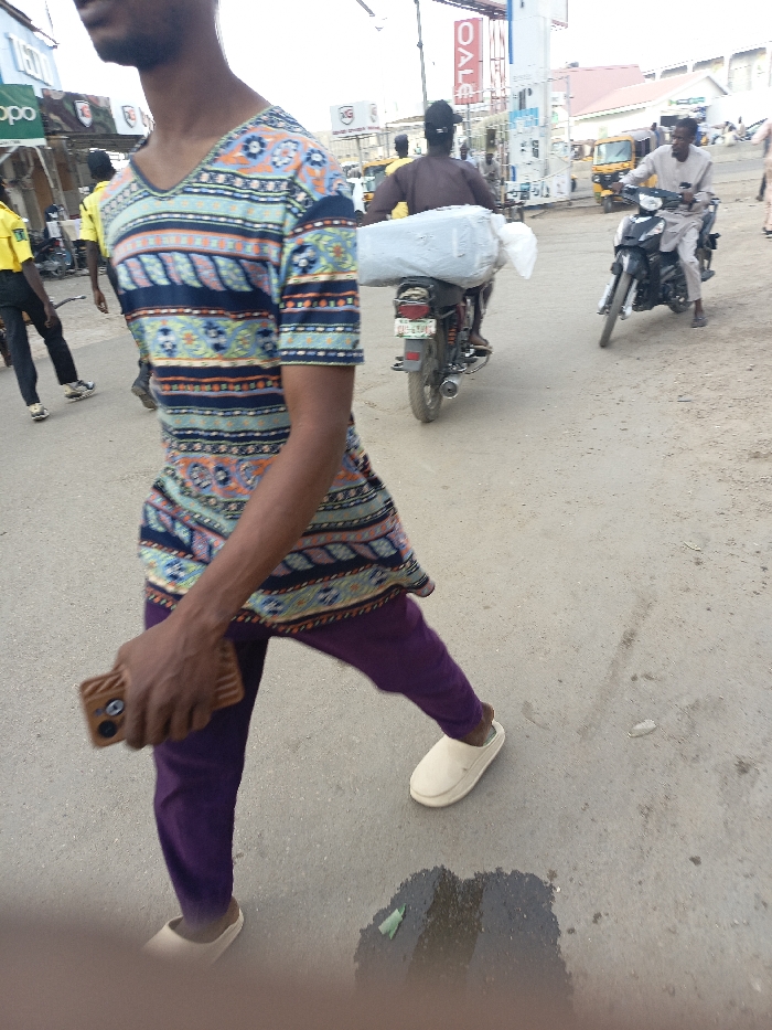 The image depicts an urban street scene in Fagge C, Nigeria, during daylight. In the foreground, partially visible from the chest down on the left, is a dark-skinned male subject walking towards the right. He wears a short-sleeved, V-neck tunic with blue, orange, and white geometric and floral patterns, purple trousers, and beige slip-on shoes. He holds a brown smartphone with a prominent camera module in his right hand. The road surface beneath him is asphalt with patches of dirt, and a wet spot with a green wrapper is visible.
In the midground, a dark motorcycle travels away from the camera, carrying a large white, possibly fabric-wrapped bundle secured to its rear rack; the rider wears a dark shirt. To its right, another dark motorcycle approaches from the background, ridden by a person wearing a light-colored, long-sleeved garment.
The background features multiple commercial buildings. On the left, signage for "OPPO" and "KGS" is visible. On the right, a vertical red sign displays the word "SALE" in white letters, adjacent to a white utility structure, possibly a fuel pump. A yellow auto-rickshaw is discernible further back on the right. Other indistinct human figures are present throughout the background. The sky appears bright and overcast.