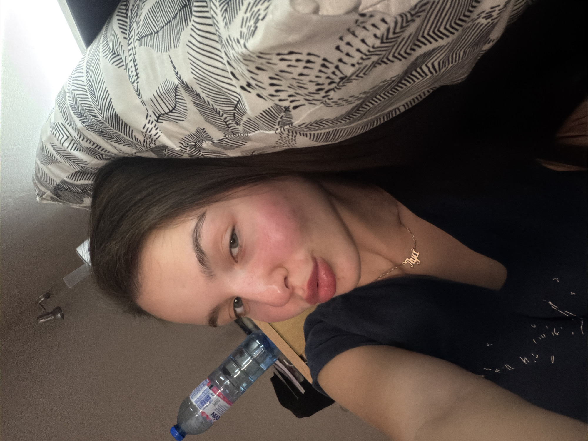 A close-up selfie of a woman lying on a pillow with a black and white leaf pattern. She has fair skin, brown hair, and a gold necklace with the name "Aya" on it. She is wearing a dark blue t-shirt. A plastic water bottle with a blue cap is visible near her face. The background is a beige-colored wall and a portion of a white fixture is visible in the distance. The scene appears to be indoors. Location is Unknown, Unknown.