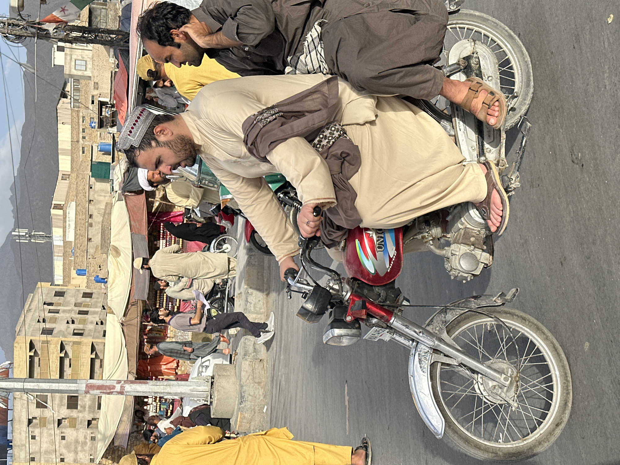 A medium shot depicts two men on a motorcycle in Quetta City Tehsil, Pakistan. The driver, wearing a cream-colored robe and a decorated cap, has a grey scarf draped around his shoulders. He wears brown sandals. The passenger behind him is dressed in brown clothing and brown sandals, with his hand near his face. The motorcycle has a red fuel tank and a silver front fender. In the background, there are buildings, tents, and other people. Some individuals are wearing traditional clothing, and there are flags visible. The overall scene suggests a busy street or market area. Mountains are visible in the distance.