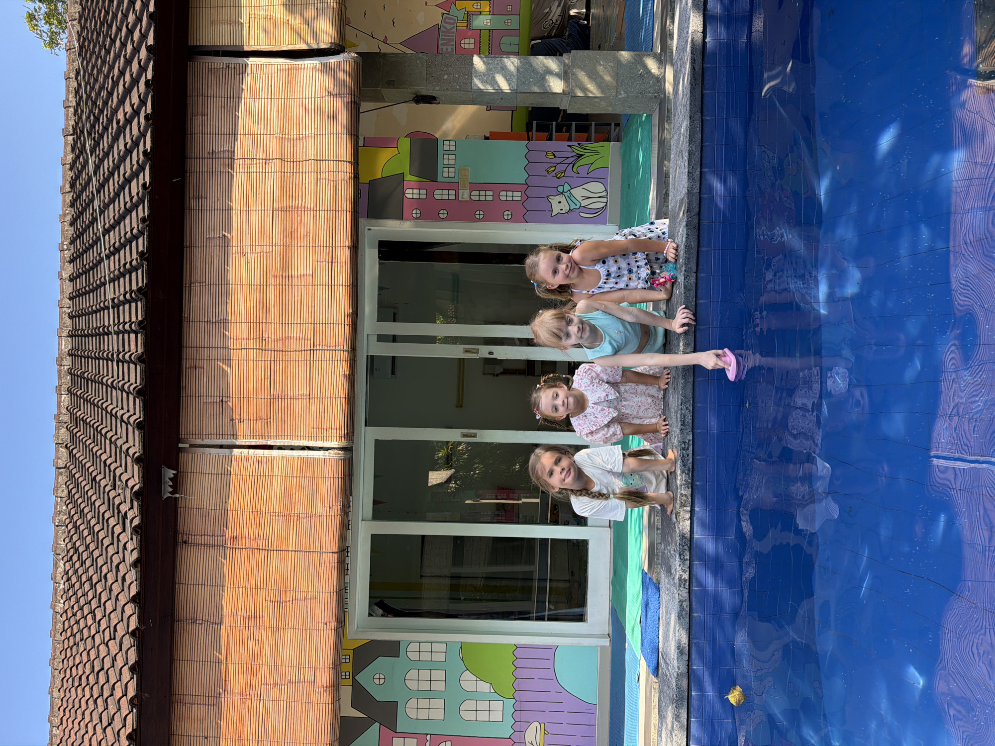 Four young girls are at the edge of a swimming pool in Kuta, Indonesia. The girls are looking at the camera. One is holding a pink toy in her hand over the water. The building behind them has a mural of colorful houses on the lower section of the exterior wall and a brown tiled roof overhead. A tan, bamboo-like material covers the upper portion of the building. The pool has blue tiled lining.