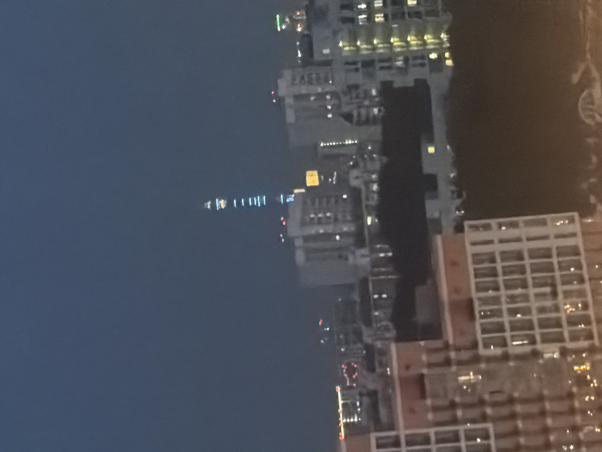 The image shows a nighttime view of a city skyline, likely Moscow, Russia. In the distance, a tall, illuminated tower stands out, with blue and white lights running vertically along its structure. Several other buildings of varying heights are visible, some with lit windows suggesting occupancy. The foreground is dominated by a large, modern building with a grid-like facade and lit windows, partially obscuring the view of other structures. The sky is a dark, dusky blue, indicating either late evening or early morning. The overall scene depicts an urban environment at night, characterized by artificial lighting and architectural structures.