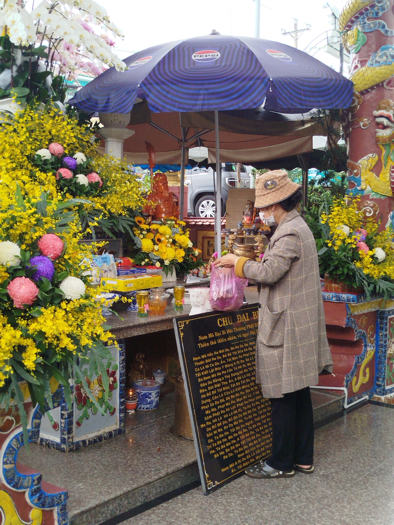 The image captures a spiritual scene at what appears to be an outdoor temple or shrine in Da Lat, Vietnam, during daytime. The central subject is a person, likely an older woman, standing in profile, facing an elaborately decorated altar. She wears a plaid long coat, black pants, a white face mask, and a brown bucket hat with a "GUCCI" logo. She is engaged in an act of devotion, holding a pink plastic bag, possibly placing or arranging offerings on the altar.
The altar is adorned with numerous vibrant flowers, including large arrangements of bright yellow blossoms (possibly Oncidium orchids), white and pink Phalaenopsis orchids, and clustered round pink, purple, and white flowers. On the altar, a reddish-brown statue (possibly a deity or Buddha) is visible, surrounded by various offerings such as packaged goods, small burning lamps, and glasses containing yellowish liquid.
A large black sign with white Vietnamese text stands prominently beside the worshipper. The text includes "CHỦ ĐẠI BỊ" and what appears to be a Buddhist mantra or prayer, specifically parts of the "Nam Mô Đại Bi Hội Thượng Phật Bản" (Great Compassion Mantra). Above the scene, a blue and purple umbrella with the "PEPSI" logo provides shade, blending modern commercial elements with the traditional setting. To the right, decorative pillars with colorful dragon motifs enhance the temple's architectural style. In the background, a silver car is partially visible. The overall atmosphere suggests reverence and cultural significance.