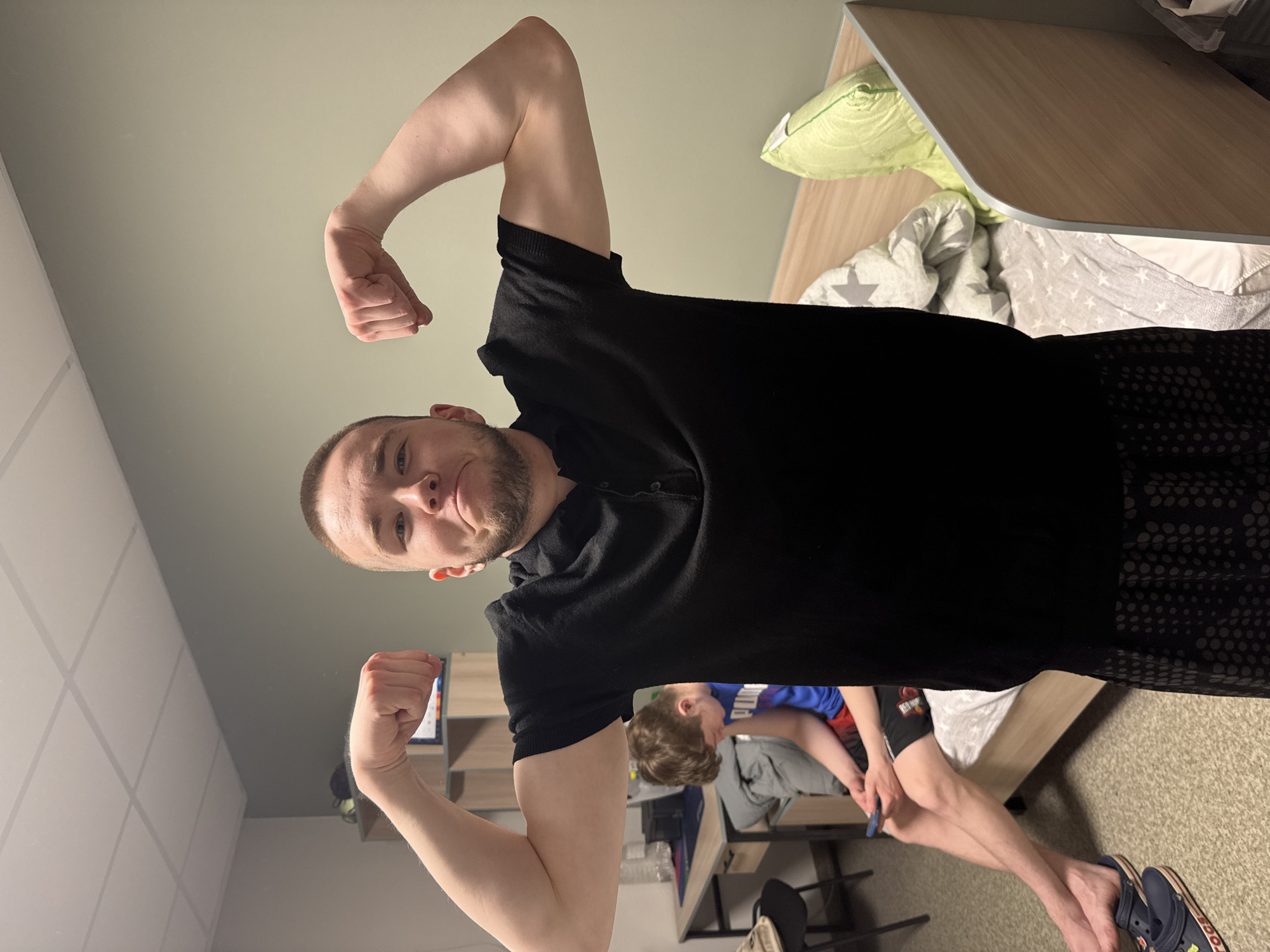 An image captured in Kazan, Russia, features a young man in the foreground, looking directly at the camera with a slight smile while flexing both biceps. He wears a black polo shirt and dark, patterned shorts. The perspective is low-angle, looking up at him.
In the background, a younger individual, possibly a child or teenager, is partially visible, seated at a desk or table. This person wears a blue and red top and shorts, with dark blue Crocs on their feet, and appears to be occupied with an object in their hands.
The indoor setting appears to be a bedroom or dormitory. Elements visible include a bed with a light green pillow and a grey duvet patterned with white stars, a light wooden headboard or shelf, and a ceiling with square white panels. Another desk or table and a chair are visible in the background. The floor is carpeted in a light shade. The lighting suggests an indoor, possibly artificial, environment, and no specific time of day or weather conditions are discernible. No visible text is present.