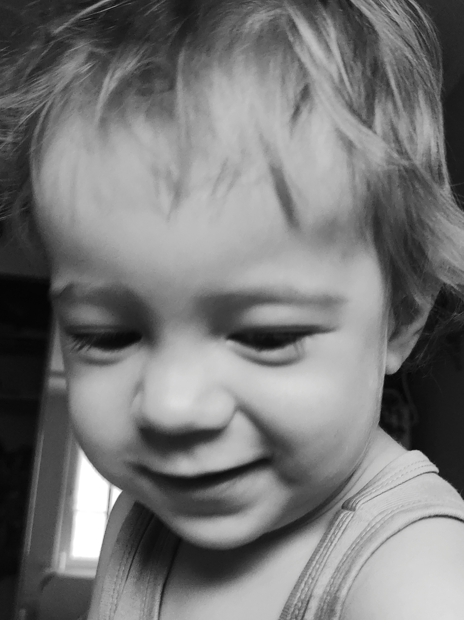 The image is a close-up, black and white portrait of a young child's face, likely taken indoors. The child, with fair, tousled hair, is looking down and slightly to the side with a gentle smile. The lighting suggests an indoor setting with a light source to the left of the frame, possibly a window. The child is wearing a tank top, the straps of which are visible. The background is dark and indistinct, with a faint hint of a window frame on the left. There is no identifiable information in the image that would allow for pinpointing a specific location within Chaikovsky, Russia.