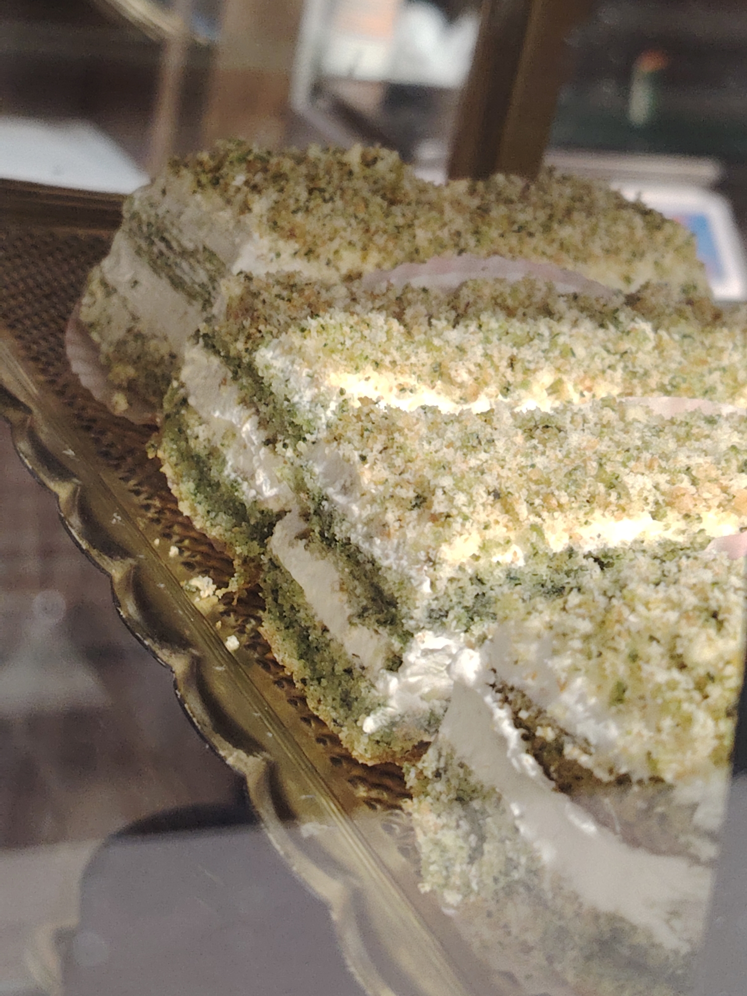 The image displays a multi-layered cake, likely a pistachio cake, presented on a gold-colored, scalloped serving tray with a textured surface, inside a glass display case within a commercial establishment in Chaikovsky, Russia. The cake consists of alternating layers of white cream or frosting and green-tinted cake, generously coated with a mixture of light green and beige crumbs. At least two slices appear to have been cut or removed from the cake. Bright overhead lighting illuminates the cake, creating reflections on the display surface below and on the glass front of the case. No individuals, actions, or interactions are visible in the scene.