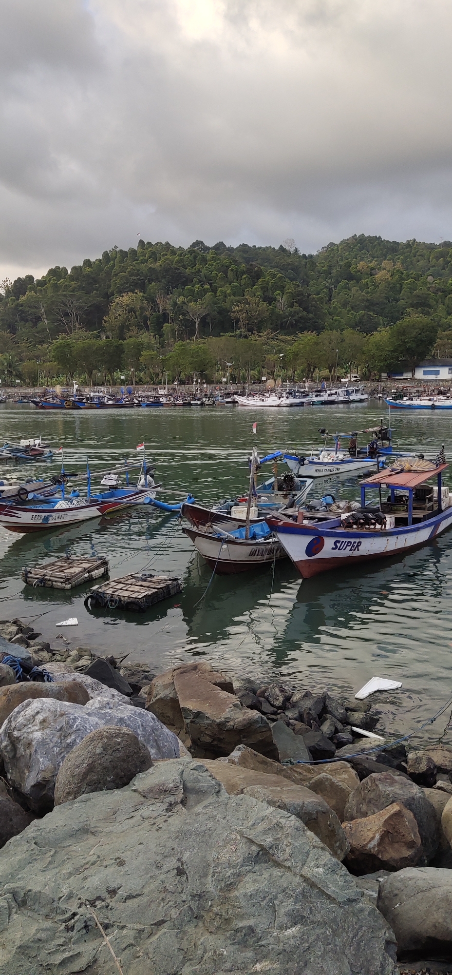 The image depicts a serene harbor scene in Tasikmadu, Indonesia, under a cloudy sky. Numerous fishing boats are anchored in the calm, green water, their colorful hulls reflecting on the surface. In the foreground, a rocky shoreline is visible, with large, rough-hewn stones forming a natural barrier. The background is dominated by a lush, tree-covered hillside, suggesting the presence of natural landscapes near the urban area. The overall atmosphere is peaceful and suggests a typical day in a coastal Indonesian fishing community.