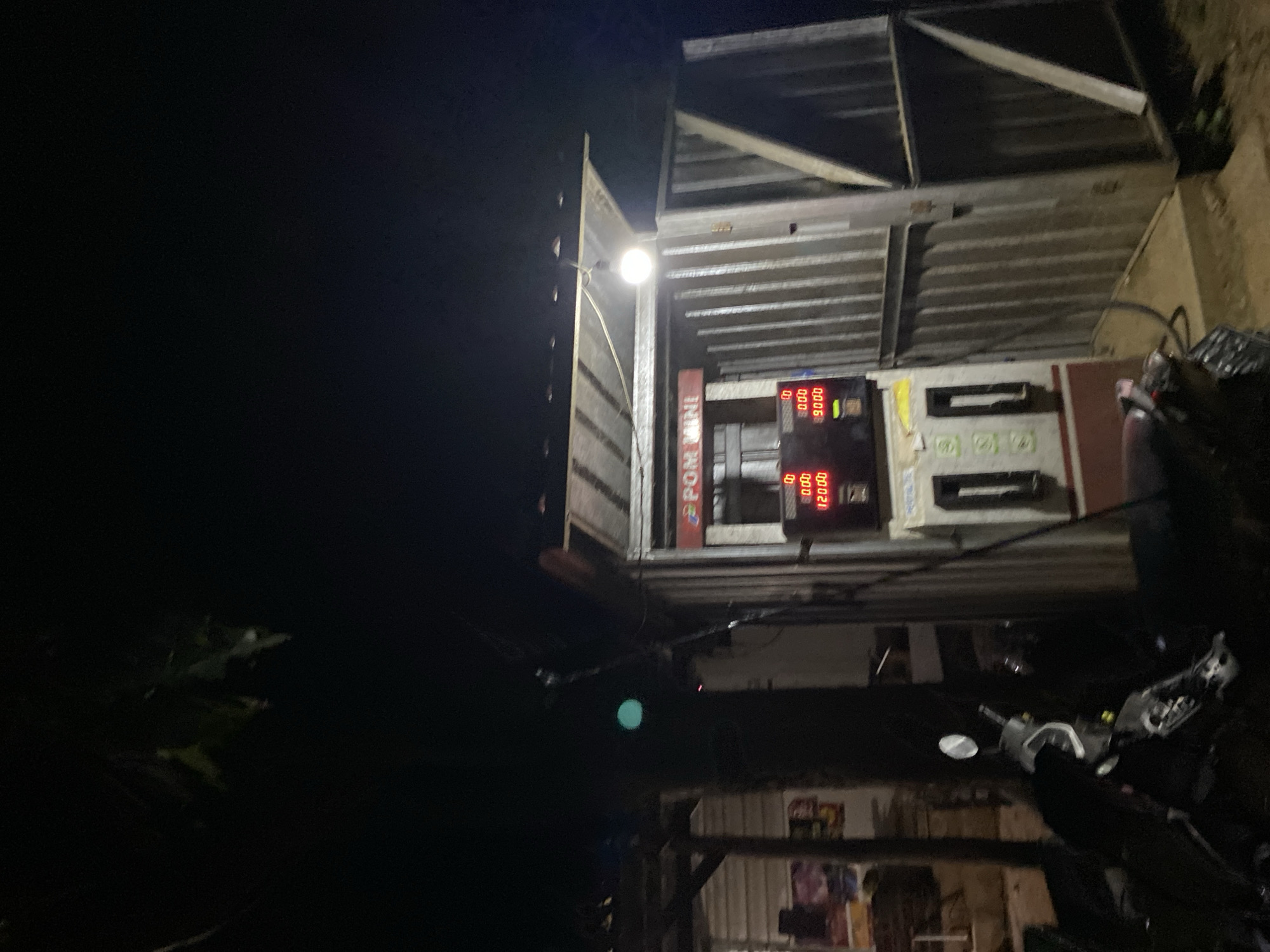 The image depicts a nighttime scene at a small, open-air gas station with a corrugated metal structure and a brightly lit gas pump. The gas pump has a digital display showing "0.00" for volume and "12000" for cost on one side, and "0.00" for volume and "9.000" for cost on the other. A sign above the pump reads "POM MINI" in red lettering. To the left of the pump, a motorcycle is parked. Behind the gas station, there appears to be a small building with a thatched or corrugated roof. The environment is dark, suggesting it is late at night, with a single light bulb illuminating the immediate area around the gas pump. The ground appears to be dirt or gravel. The background is obscured by darkness.