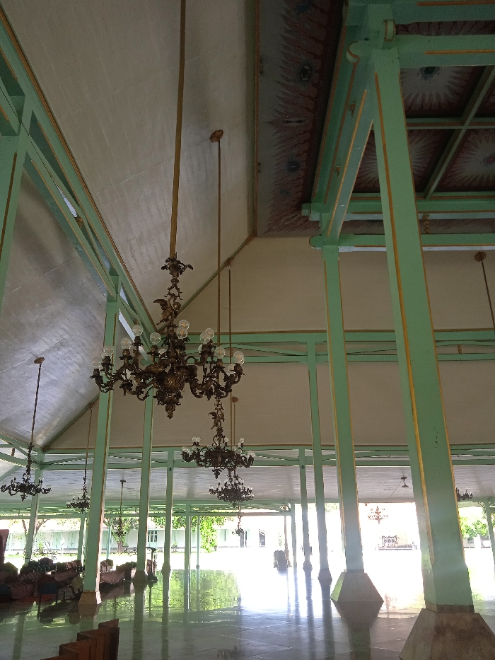 The image shows an interior view of a building. The room has a high ceiling with several large pillars supporting the roof. The ceiling is painted white with green beams. The pillars are painted light green and have gold accents. There are several ornate chandeliers hanging from the ceiling. The room is empty and appears to be a hallway or passageway. The room is well-lit, and the floor is clean and shiny. The image appears to be taken at the Surakarta Palace in Surakarta, Indonesia, located at coordinates -7.559 latitude and 110.783 longitude.