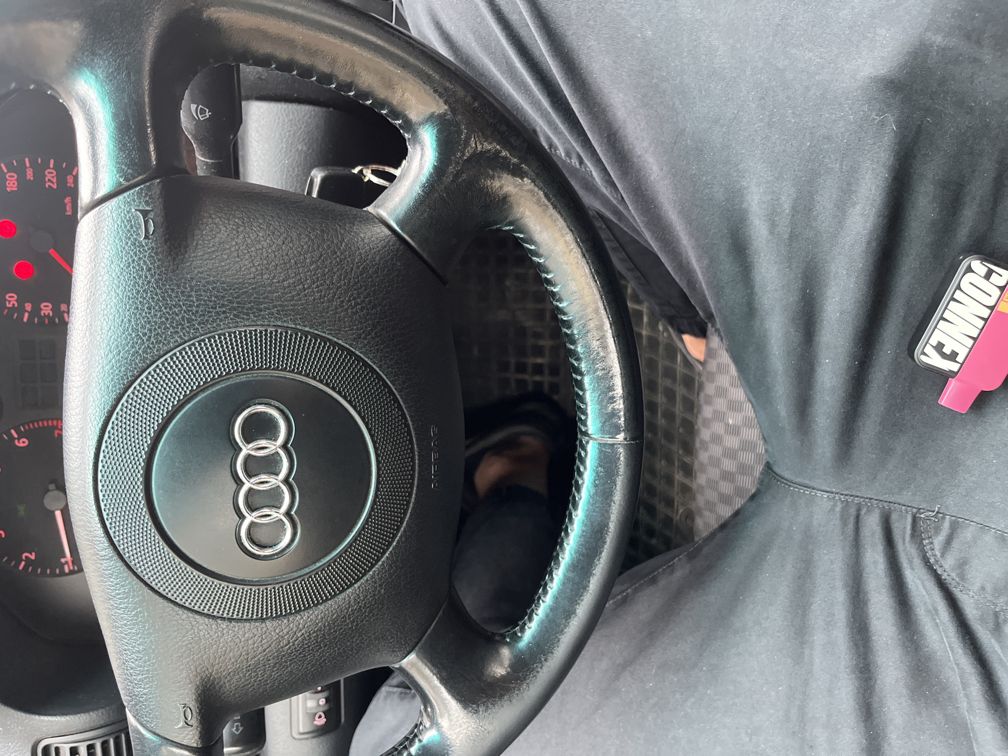 A driver is behind the wheel of an Audi, possibly in Sofia, Bulgaria. The car features a black steering wheel with the Audi logo. The driver is wearing dark gray or black clothing. The speedometer is visible. The floor of the car has a checkered pattern. A key fob or accessory with the word "CONNE" is attached to the driver's clothing near the waist.