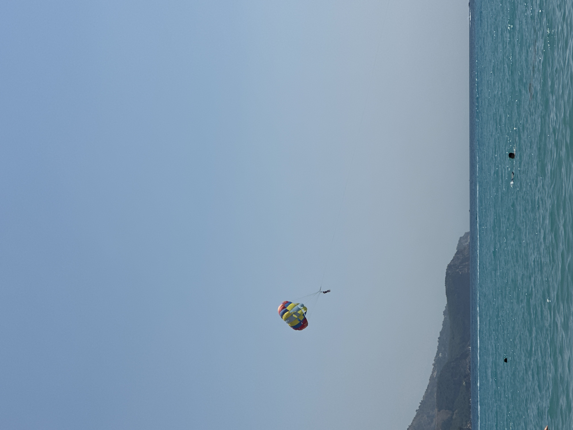 In the city of Rafailovići, Montenegro, a vibrant parasailer with a colorful parachute soars high above the tranquil turquoise waters of the Adriatic Sea. The parasailer, suspended by a rope, appears as a small, dark silhouette against the expansive, clear blue sky. In the foreground, the sea is calm, with gentle ripples reflecting the sunlight. A few indistinct figures can be seen swimming or wading in the water, their dark heads barely breaking the surface. In the background, a lush, green mountain range slopes down to the coastline, providing a scenic backdrop to the coastal activity. The scene is bathed in bright, natural light, indicating a clear, sunny day.