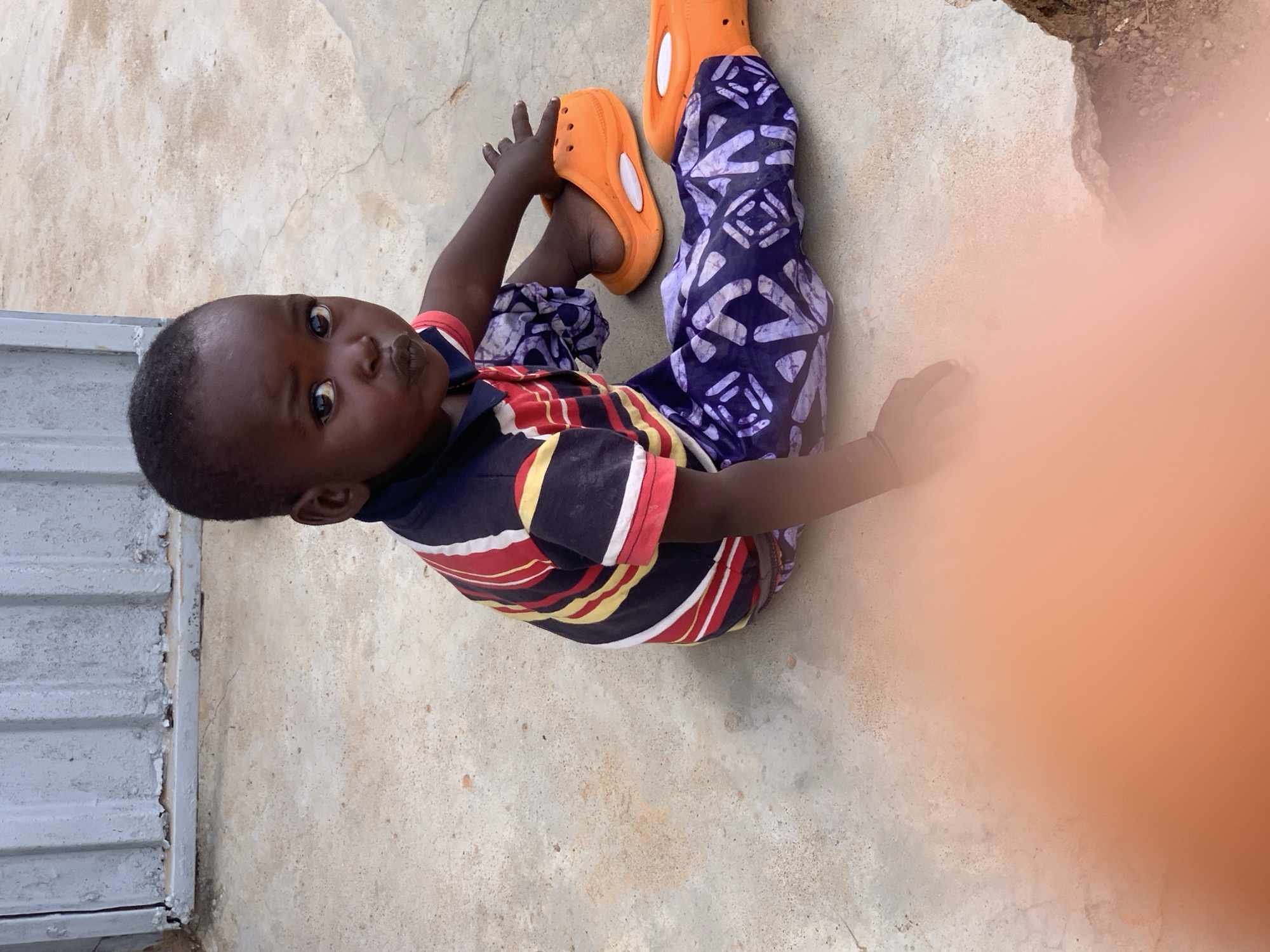 A young Black child, likely a toddler, sits on a rough concrete surface in Sabon Tasha, Nigeria. The child has dark skin, dark hair, and large, curious eyes. They are wearing a colorful striped t-shirt with red, yellow, blue, and white stripes, and dark blue pants with a white pattern. The child is also wearing bright orange slip-on shoes. Their left leg is extended in front of them, with the orange shoe visible. Their right hand rests on their right shoe, also orange. The child's mouth is pursed, giving them a slightly pouty expression. In the background, a portion of a gray metal corrugated wall or structure is visible. The concrete ground is cracked in places, and there are small pebbles and dirt scattered around. A large, out-of-focus orange area is visible in the bottom left and right of the frame, suggesting a finger or lens flare may be obscuring part of the scene.